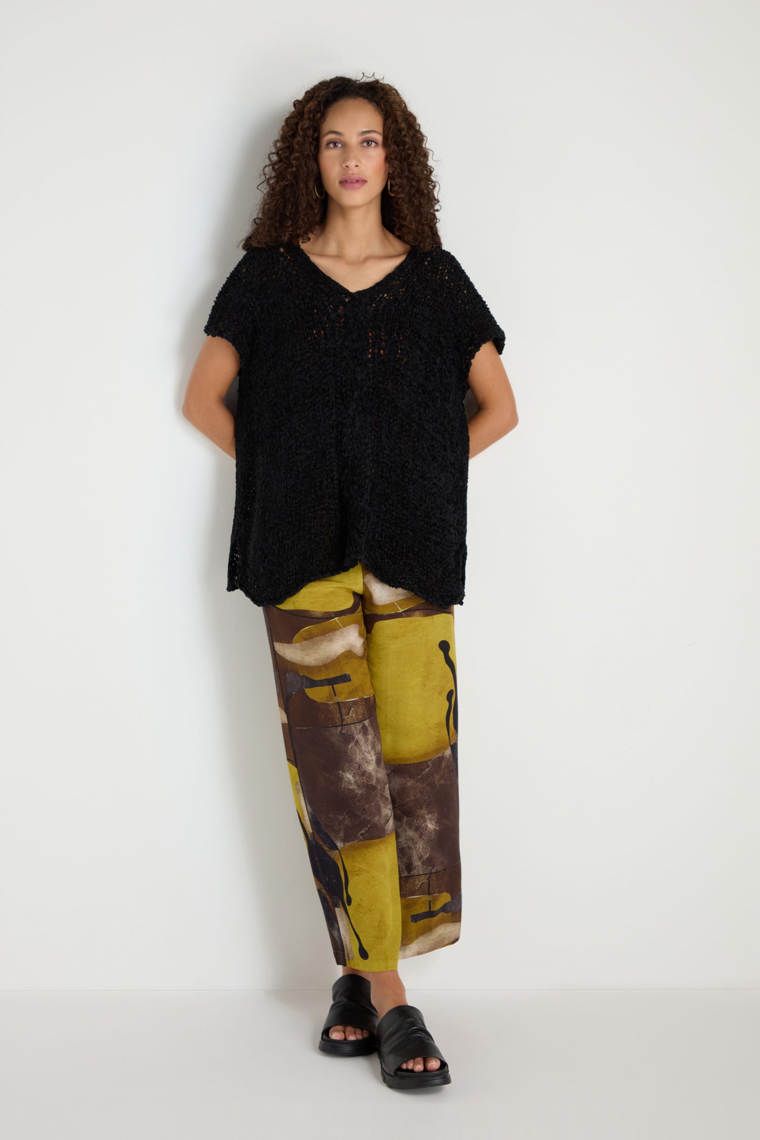 A woman stands against a white wall wearing a relaxed silhouette V-Neck Cotton Blend Black Knit, wide-leg abstract print pants in earth tones, and black slide sandals.