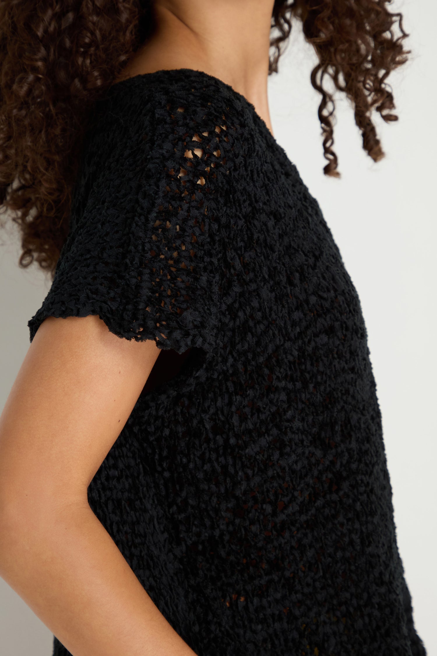Person with curly hair wearing the V-Neck Cotton Blend Black Knit top, short-sleeved and textured, shown from shoulder to mid-arm against a neutral background.