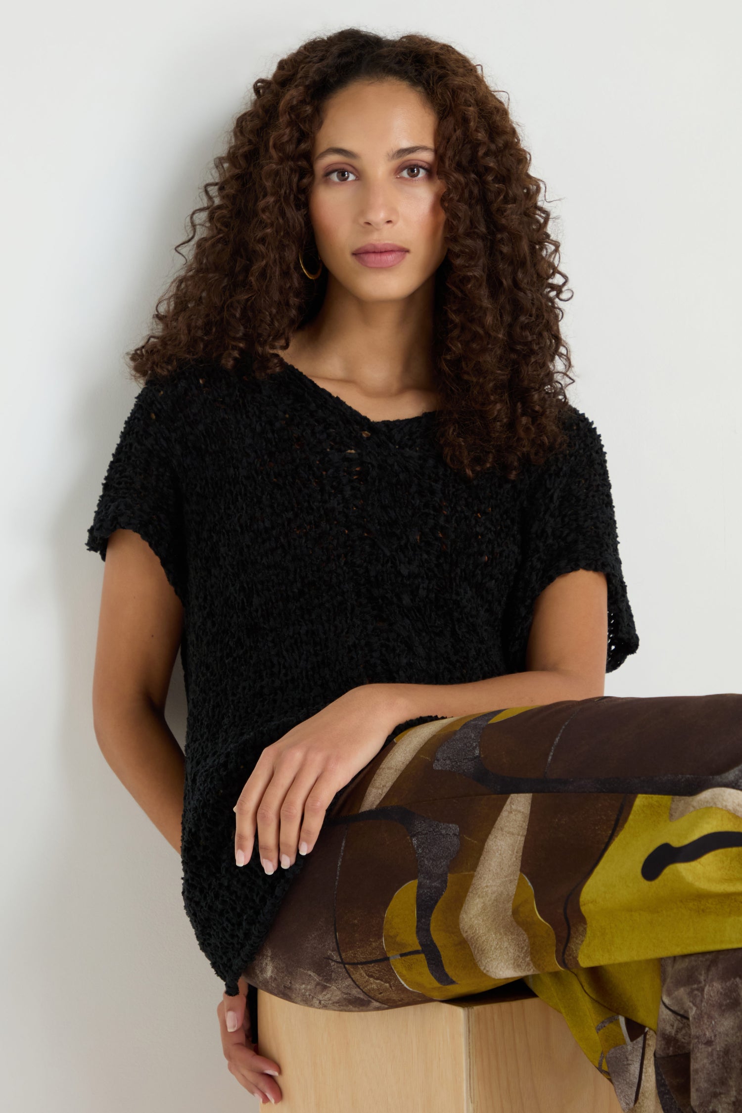 A woman with curly hair wears the V-Neck Cotton Blend Black Knit top and abstract-patterned pants, sitting on a light wooden block against a plain light background.