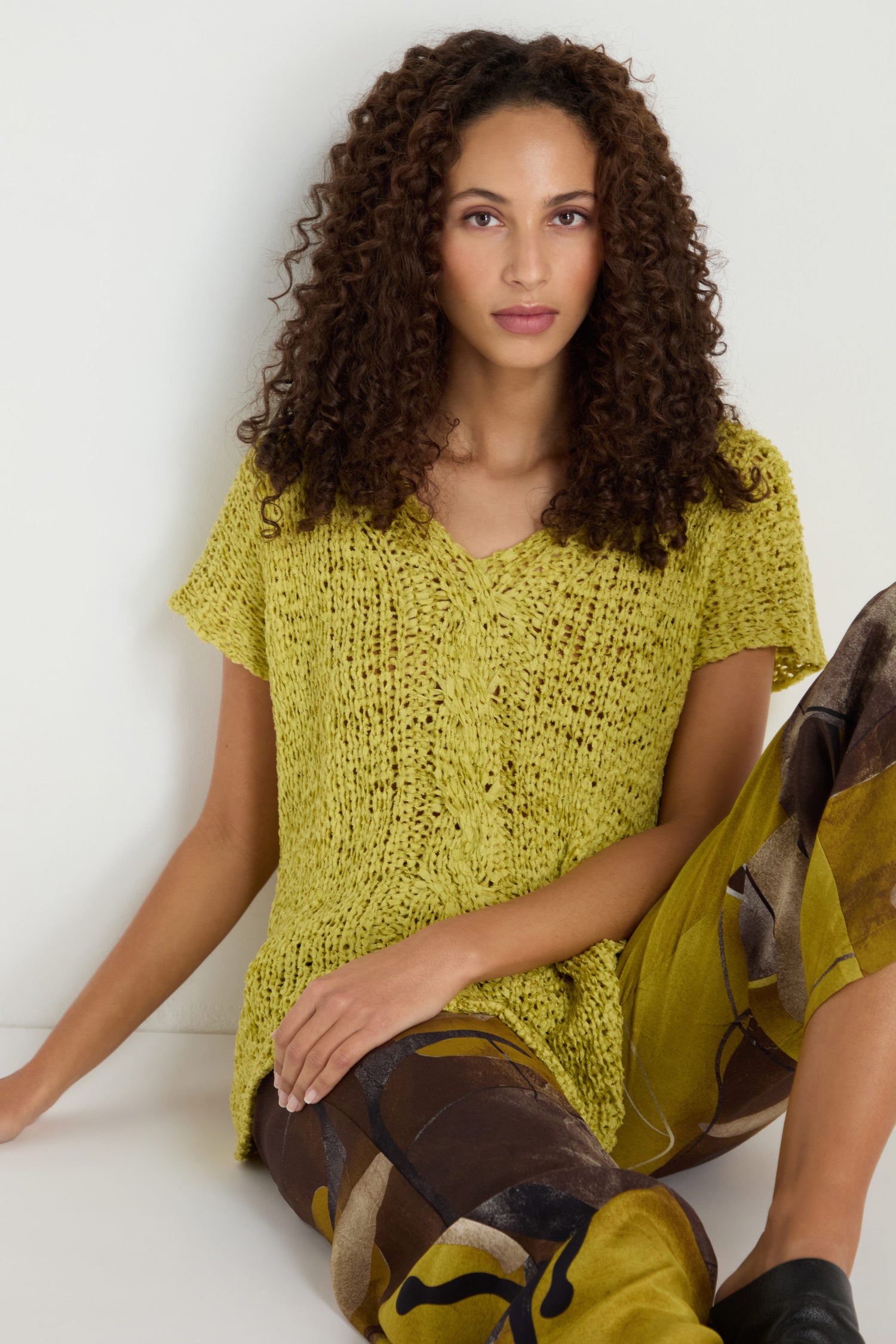 A woman with curly hair wears the V-Neck Cotton Blend Knit in yellow and patterned brown-and-yellow pants, seated against a white background.