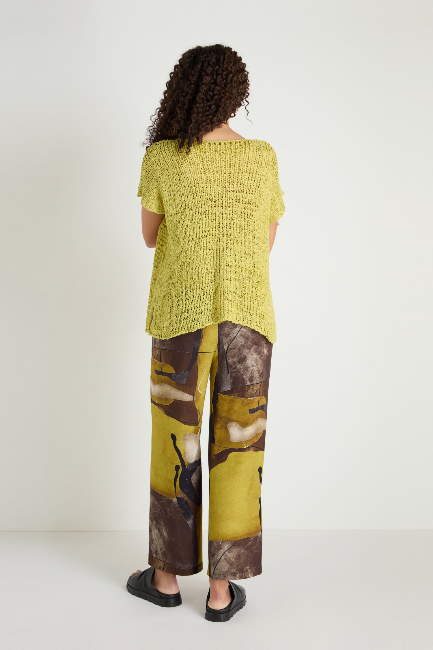 A person with curly hair faces away, wearing the V-Neck Cotton Blend Knit in yellow, abstract brown and yellow pants, and black slip-on shoes against a plain white background.