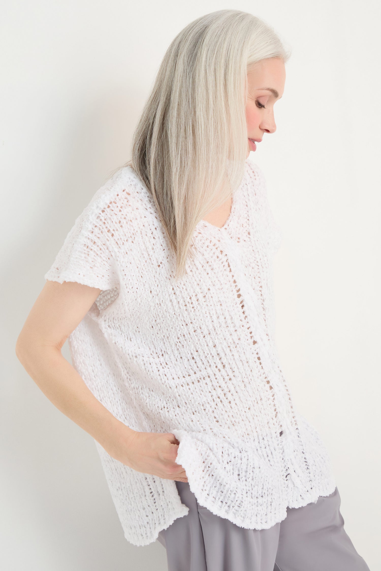 A woman with long gray hair wears the V-Neck Cotton Blend Knit and light gray pants, standing against a plain white background with one hand in her pocket, looking downward.