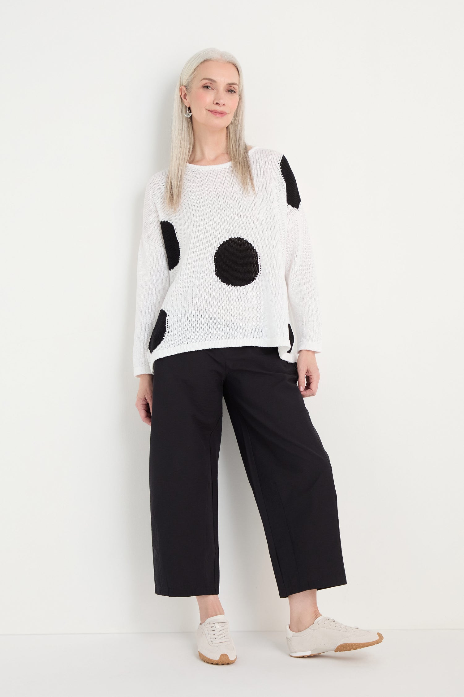 Someone stands against a white wall wearing the Spot Cotton Knit sweater, black wide-leg pants, and white sneakers for a relaxed silhouette.