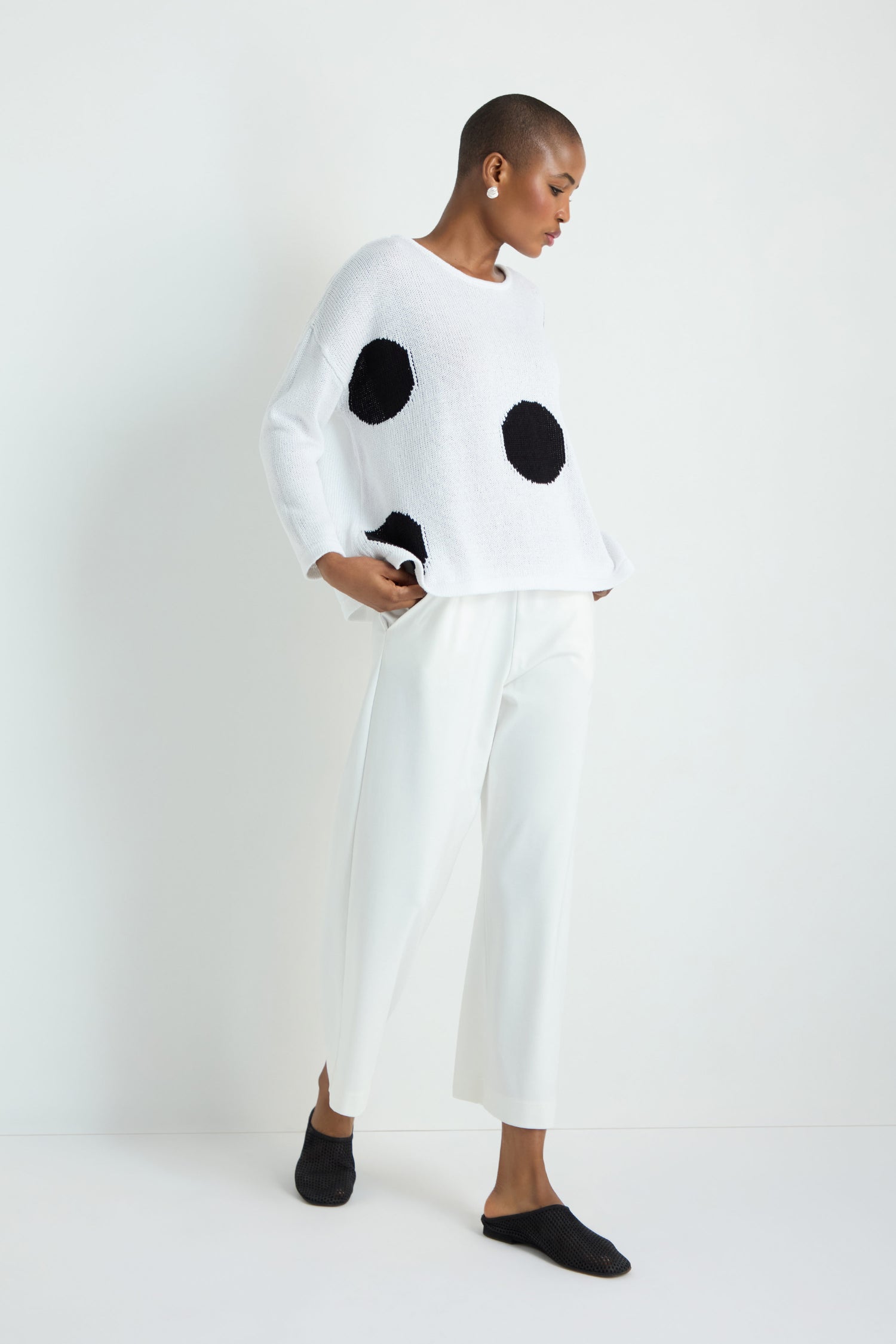 Wearing the Spot Cotton Knit sweater with bold spots, a person with long, straight, light hair pairs it with black wide-leg pants and white sneakers, standing against a plain white background.