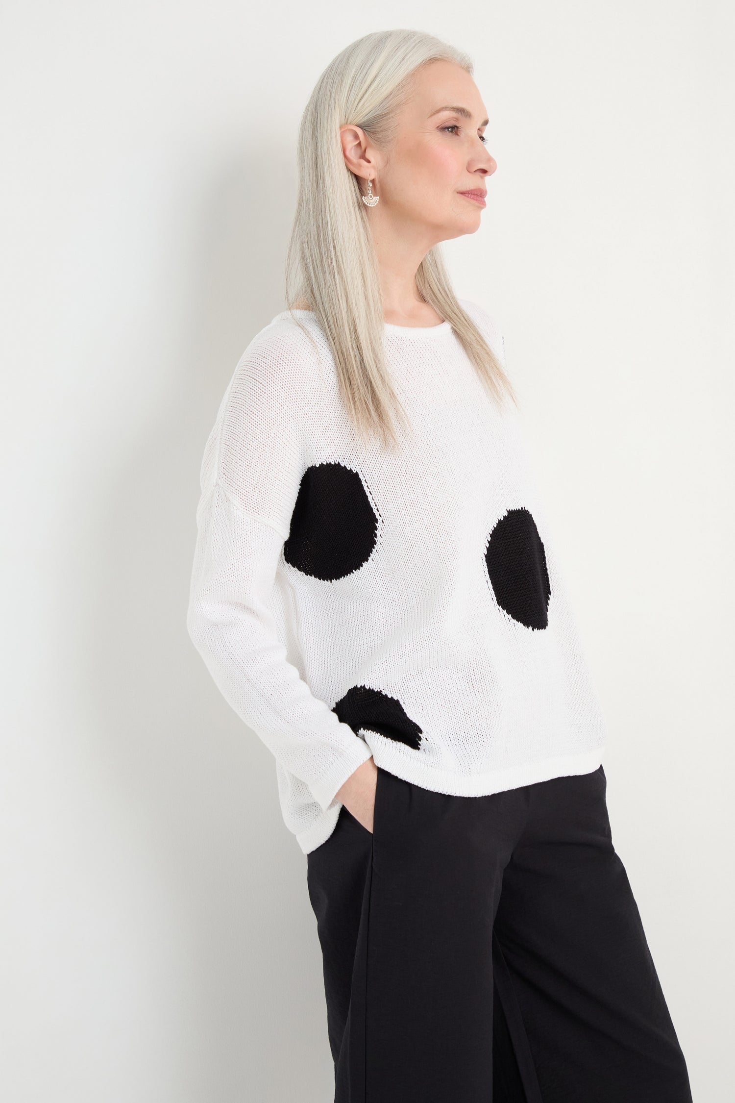 A woman with long gray hair wears the Spot Cotton Knit, a white organic cotton sweater with a bold spot pattern, paired with black pants and standing against a plain white background.