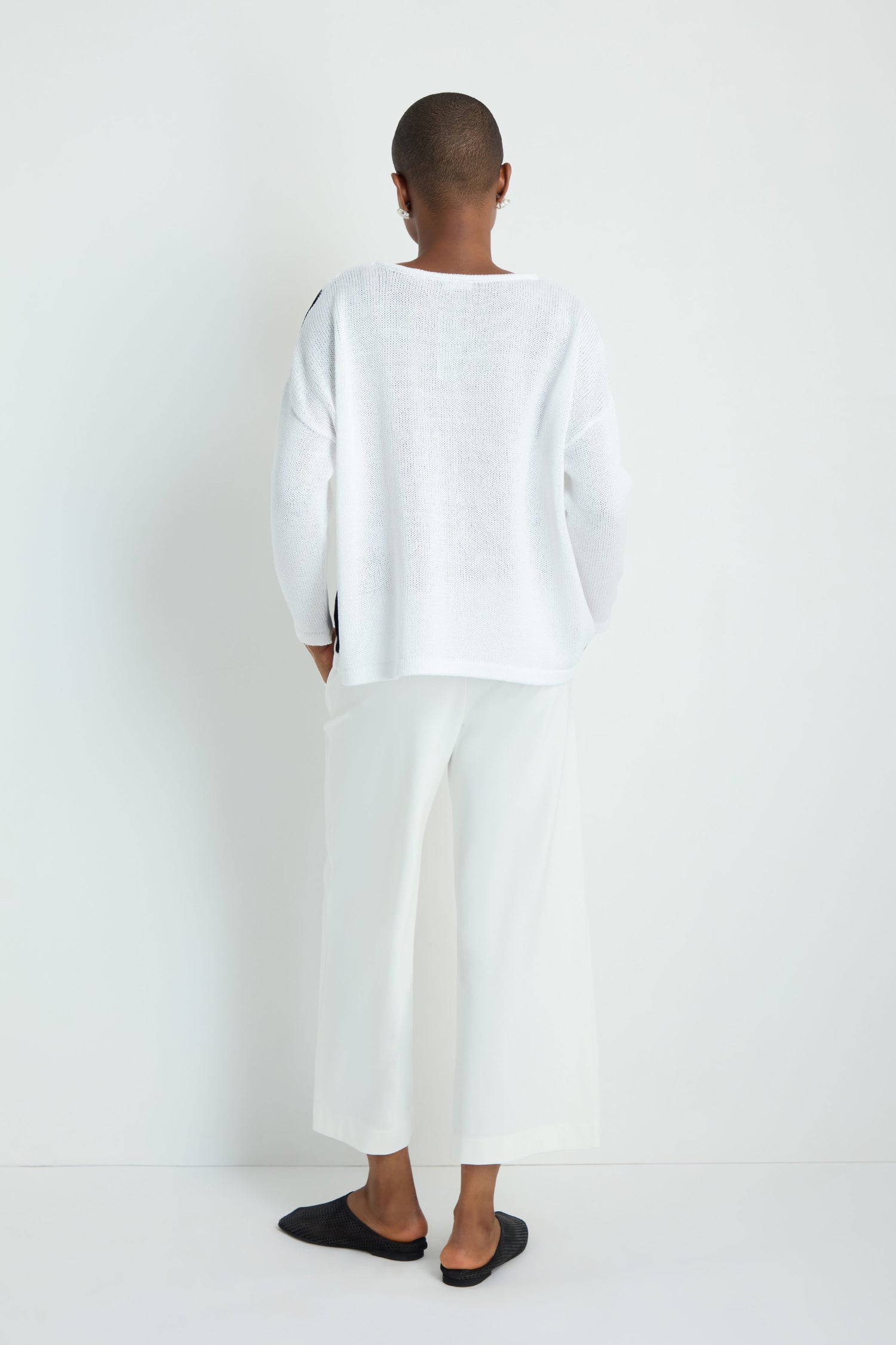Wearing the Spot Cotton Knit in white, a person with long gray hair and black wide-leg pants stands facing a white wall, back to the camera, highlighting the sweater's relaxed silhouette.