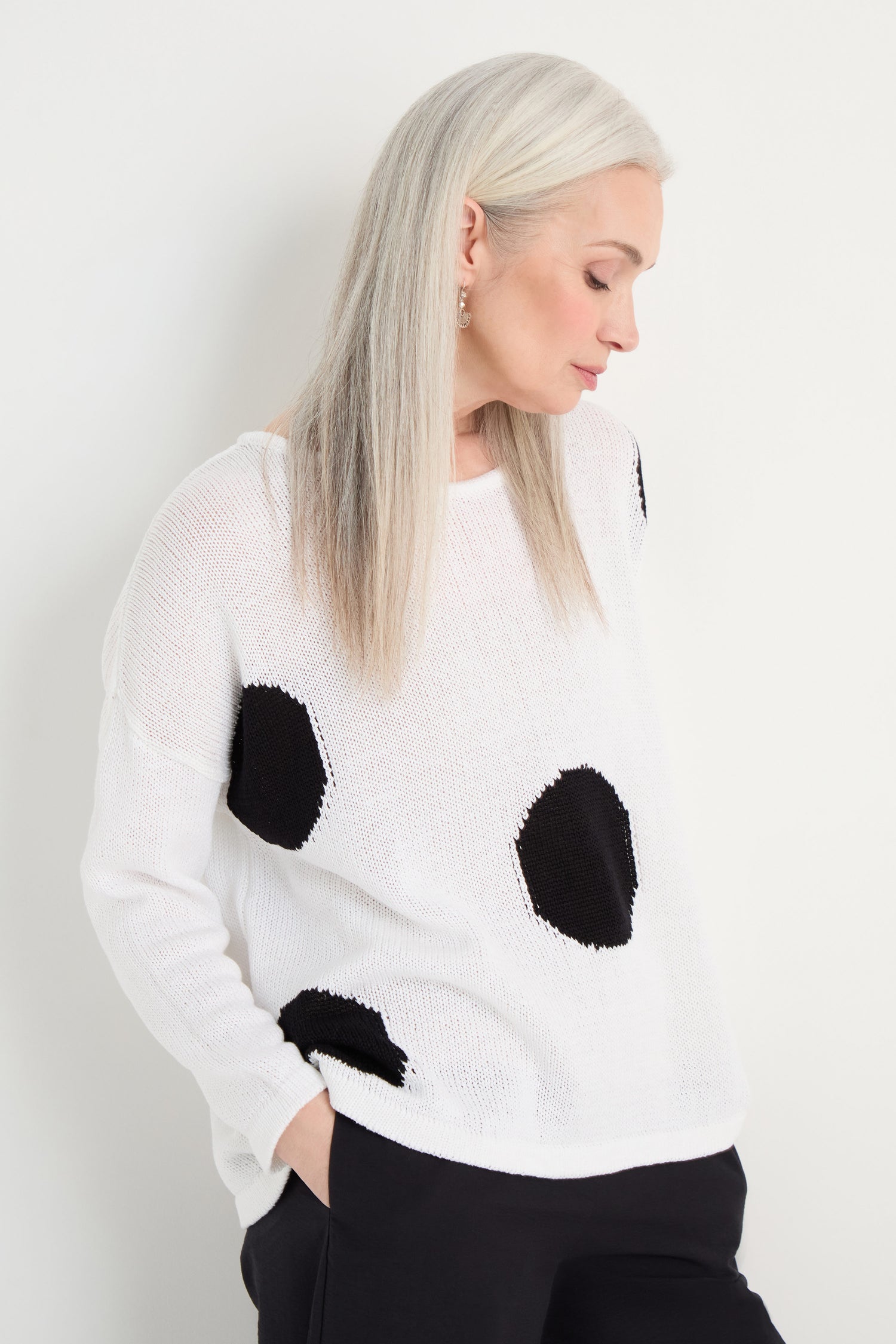 A woman with straight, long gray hair wears the Spot Cotton Knit—a white sweater with a bold spot pattern in organic cotton—and black pants, standing against a plain white background with her hands in her pockets for a relaxed silhouette.