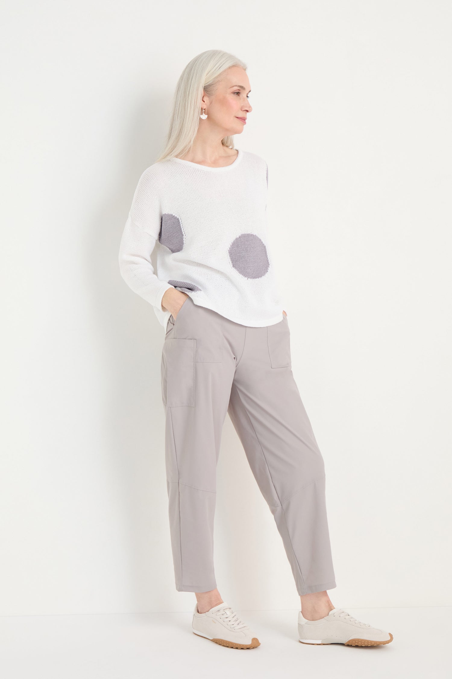 Older woman with long white hair in a relaxed silhouette wears the Spot Cotton Knit—white sweater with bold spots—paired with light gray pants and white sneakers, standing and looking to the side against a plain background.