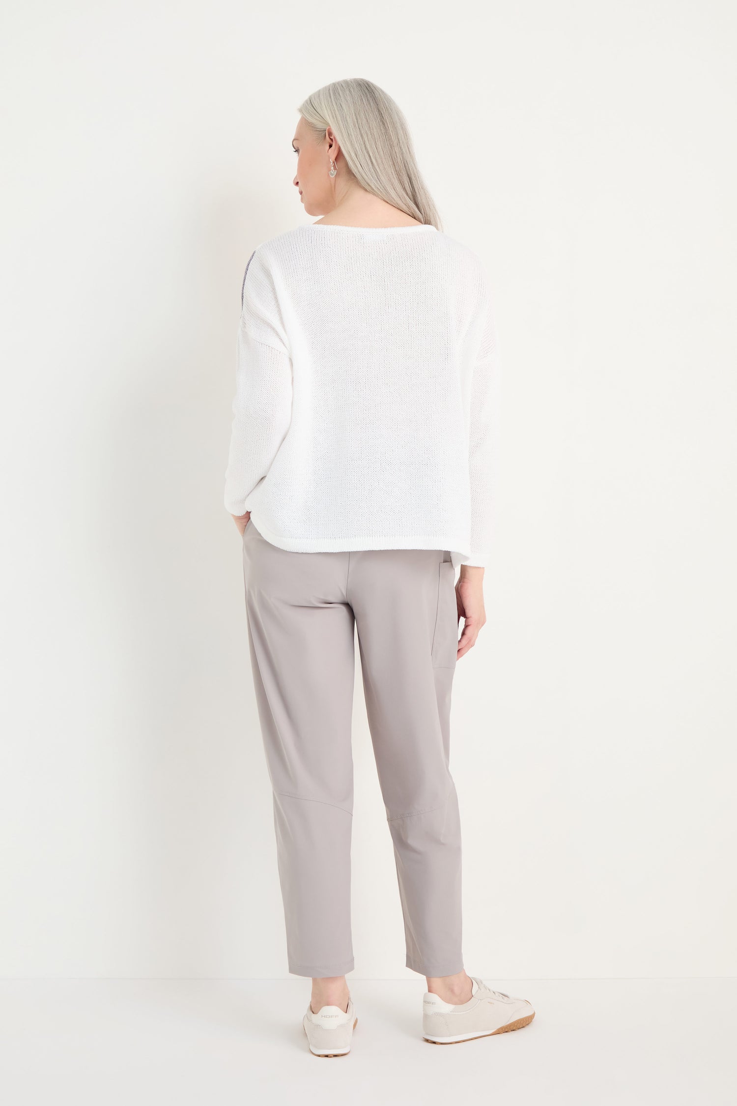 An older woman with long gray hair faces away, wearing the Spot Cotton Knit top in white, light gray pants, and white sneakers against a plain white background.