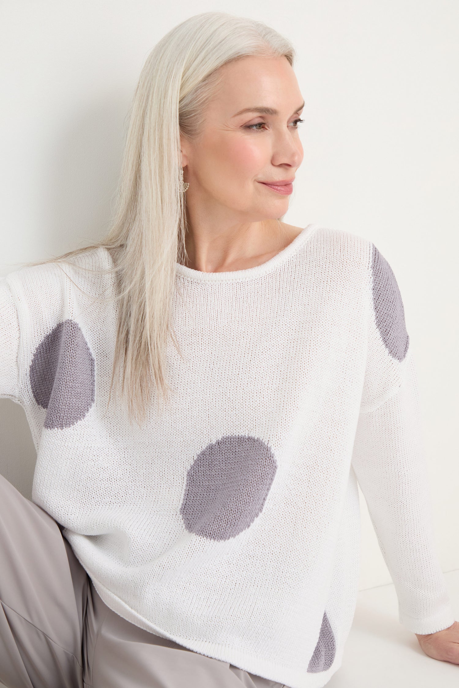A woman with long gray hair sits against a white wall, wearing the Spot Cotton Knit sweater with a bold spot pattern and light-colored pants, looking to the side and smiling slightly.