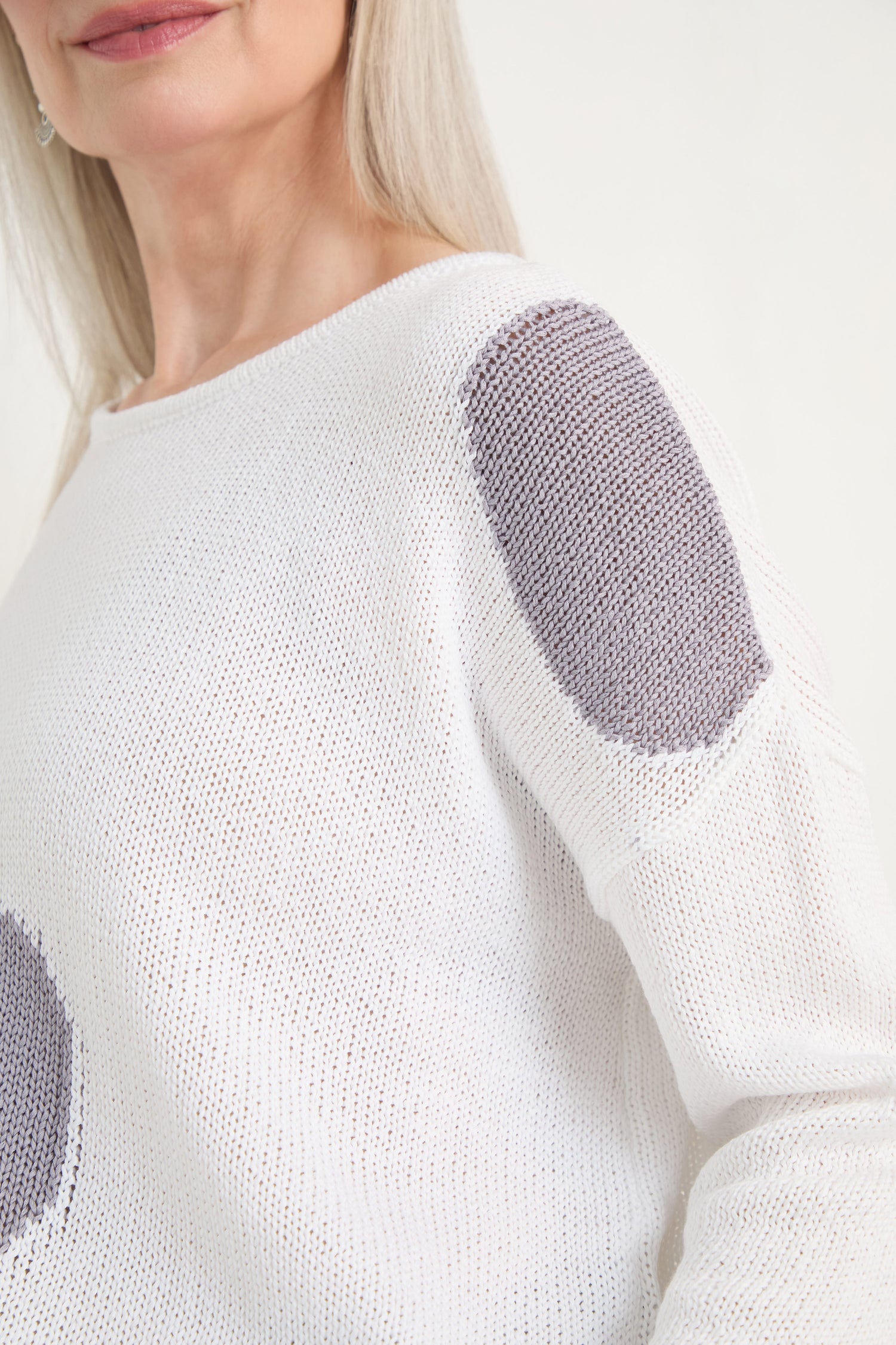 Close-up of a woman wearing the Spot Cotton Knit, an organic cotton white sweater featuring gray oval elbow patches, against a plain background.
