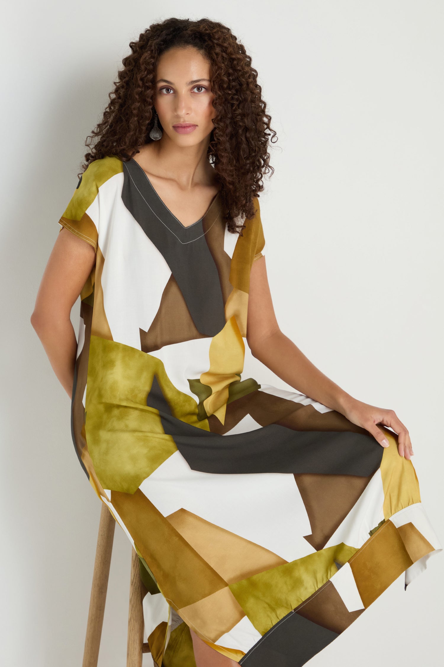 A woman with curly hair sits on a wooden stool against a neutral background, wearing a loose-fitting, short-sleeved Collage Print V-Neck Dress in green, brown, mustard, and white.