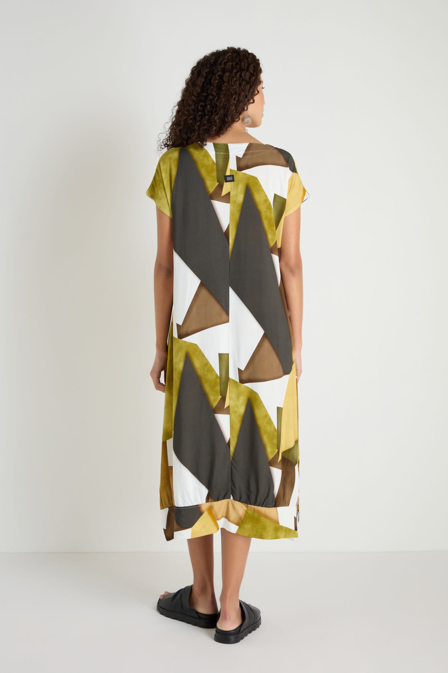 A woman stands facing away in a Collage Print V-Neck Dress featuring bold yellow, black, brown, and white geometric patterns, paired with black slip-on sandals in a plain white room.