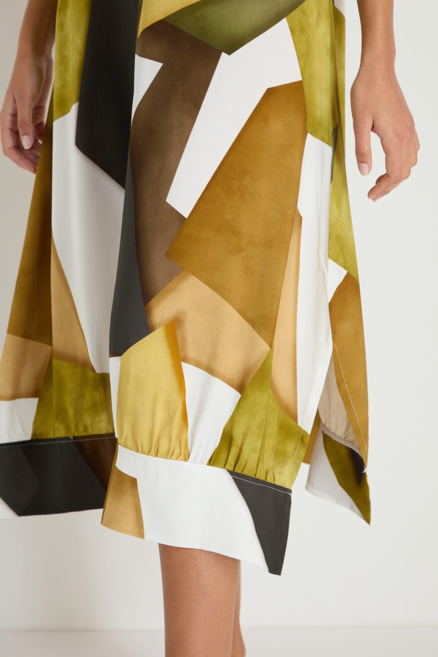 A person wears the Collage Print V-Neck Dress, featuring a geometric pattern in green, brown, white, and yellow. The dress is shown from mid-thigh to calf with one hand visible at the side.