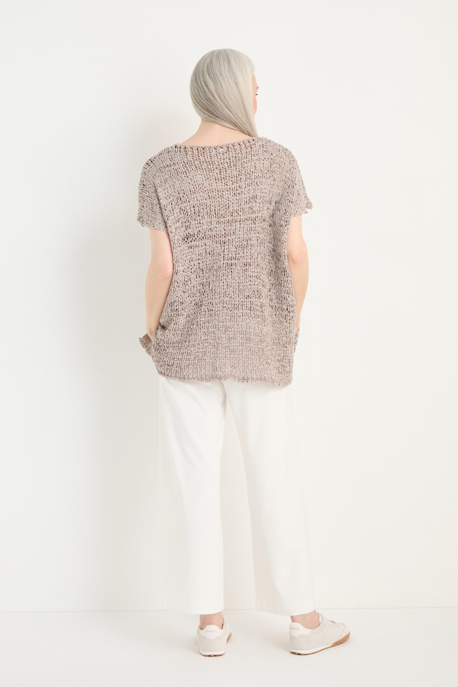 A person with long gray hair faces away, wearing a loose-knit beige top and Wide Leg Pocket Trousers in white viscose stretch with an elasticated waist, paired with white sneakers against a plain white background.