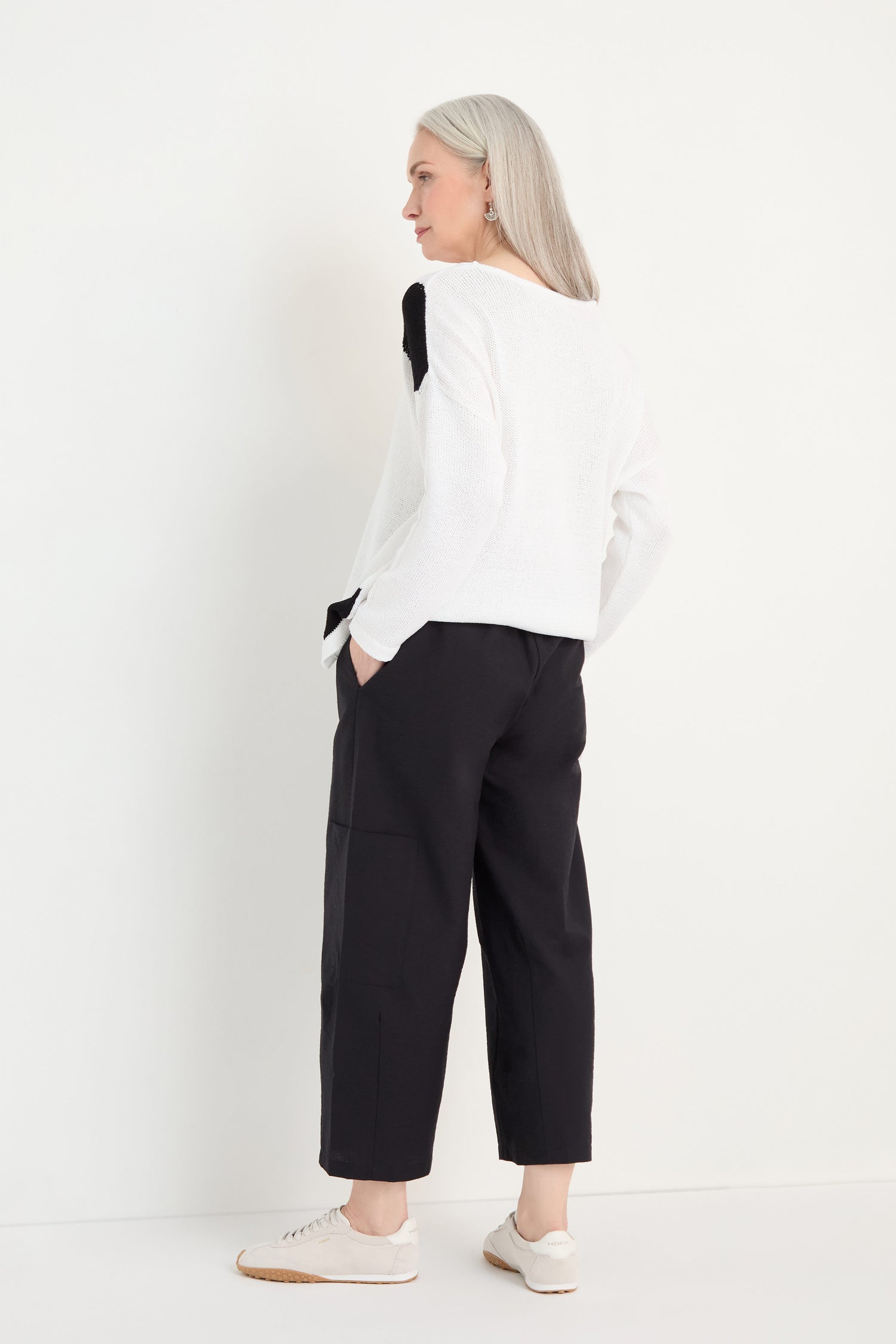 A person with long gray hair stands facing away, wearing a white sweater, Wide Leg Cotton Trousers, and white sneakers with hands in pockets against a plain white background.