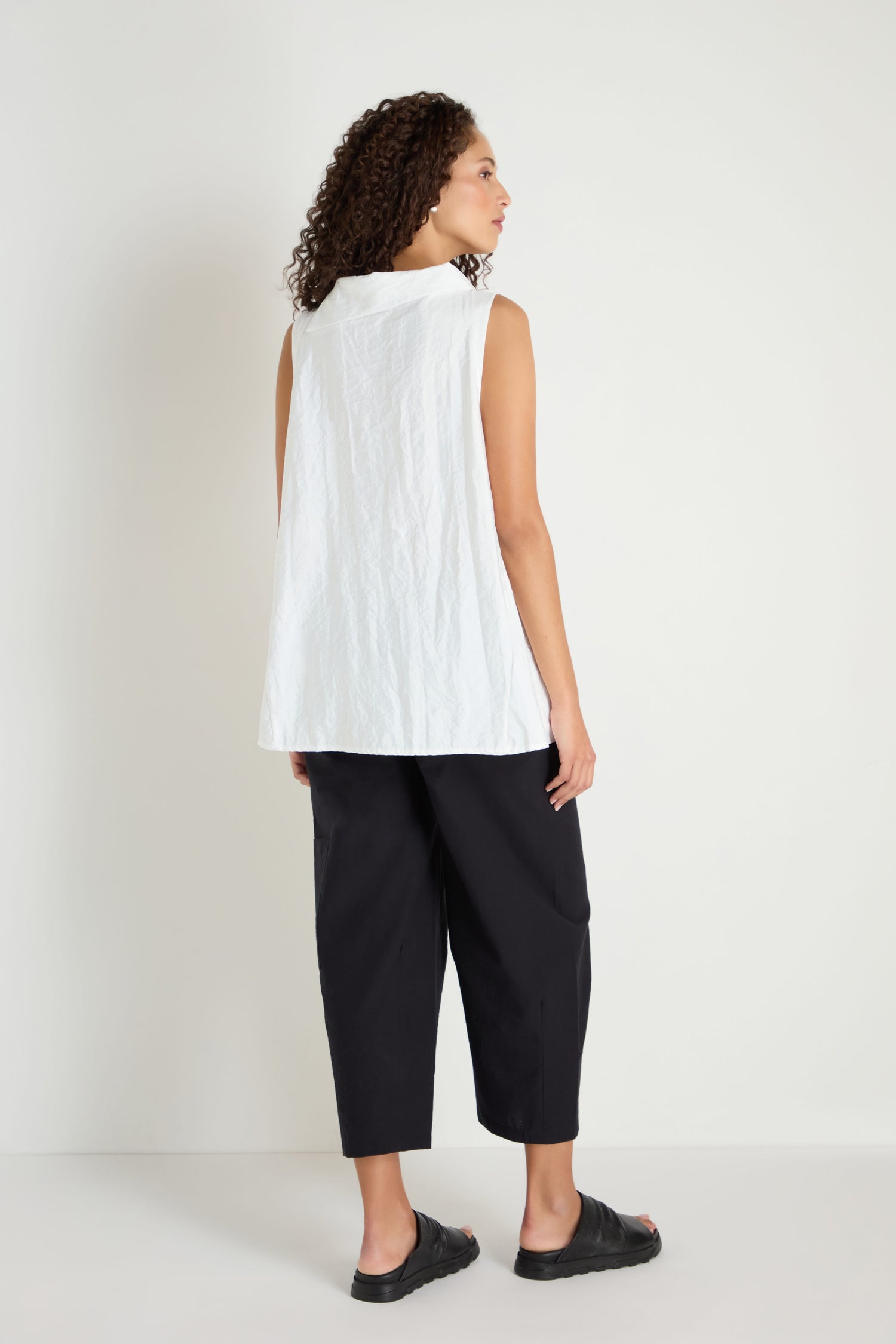 Person standing against a white wall, wearing the Roll Collar Step Hem Top, black cropped pants, and black slide sandals, facing slightly away from the camera.