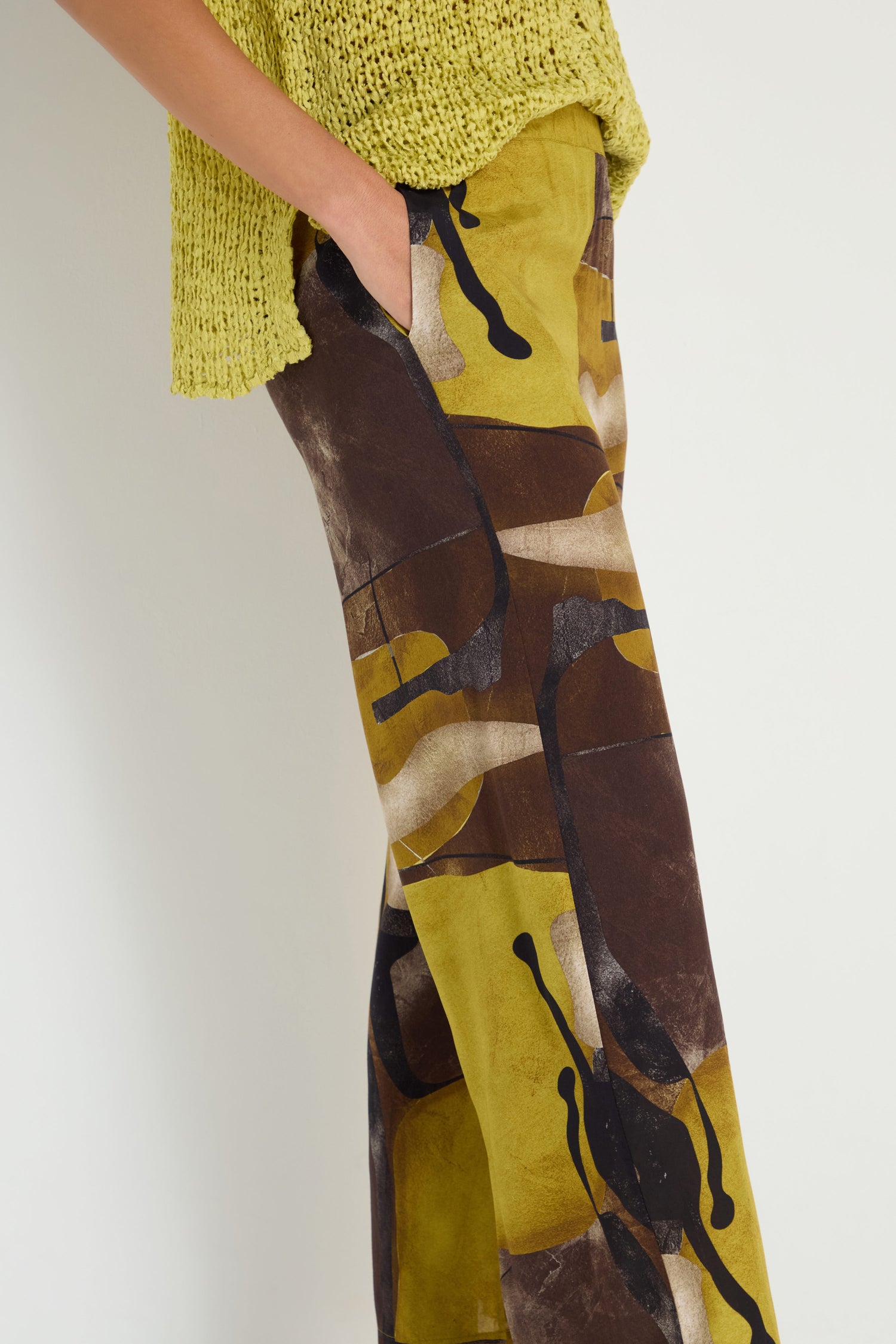 Person wears a yellow-green knit top and Abstract Print Trousers in brown, yellow, and black viscose, standing with one hand in pocket against a plain background.
