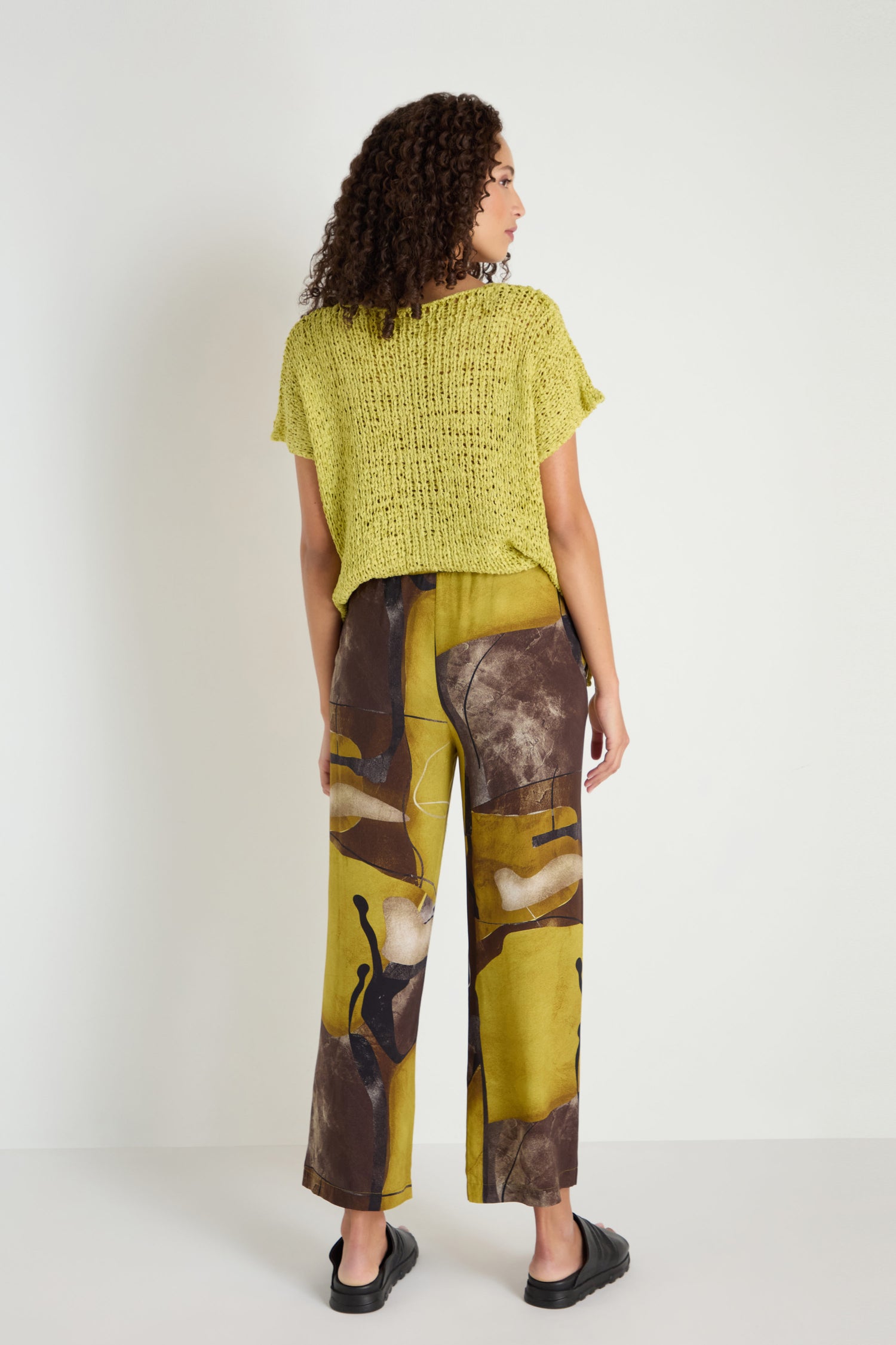 A person stands facing away, wearing a loose yellow knit top, Abstract Print Trousers in brown and yellow tones with a relaxed wide fit, and black slip-on shoes.