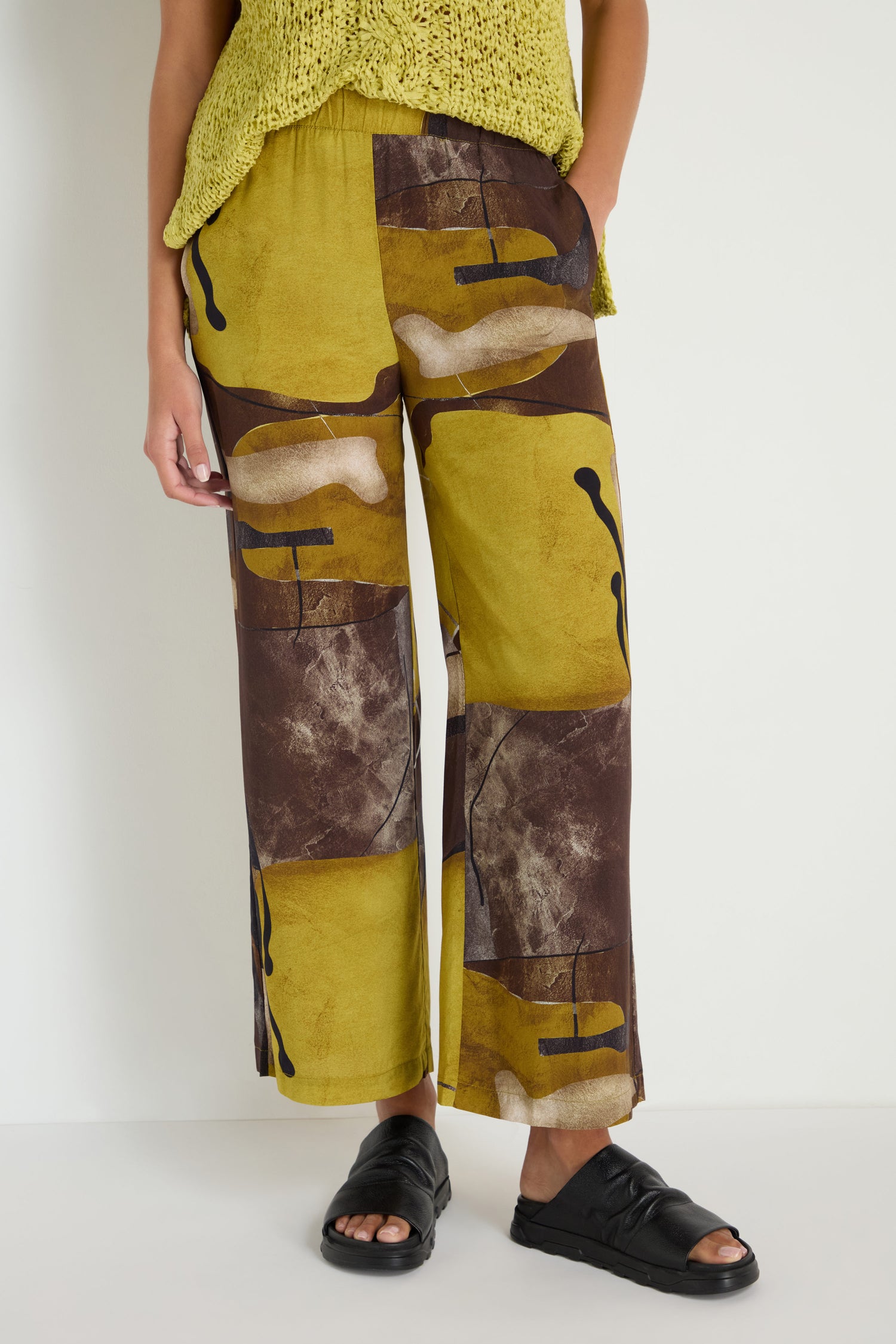 A person stands against a plain white wall wearing Abstract Print Trousers in yellow and brown, paired with a yellow knit top and black sandals.