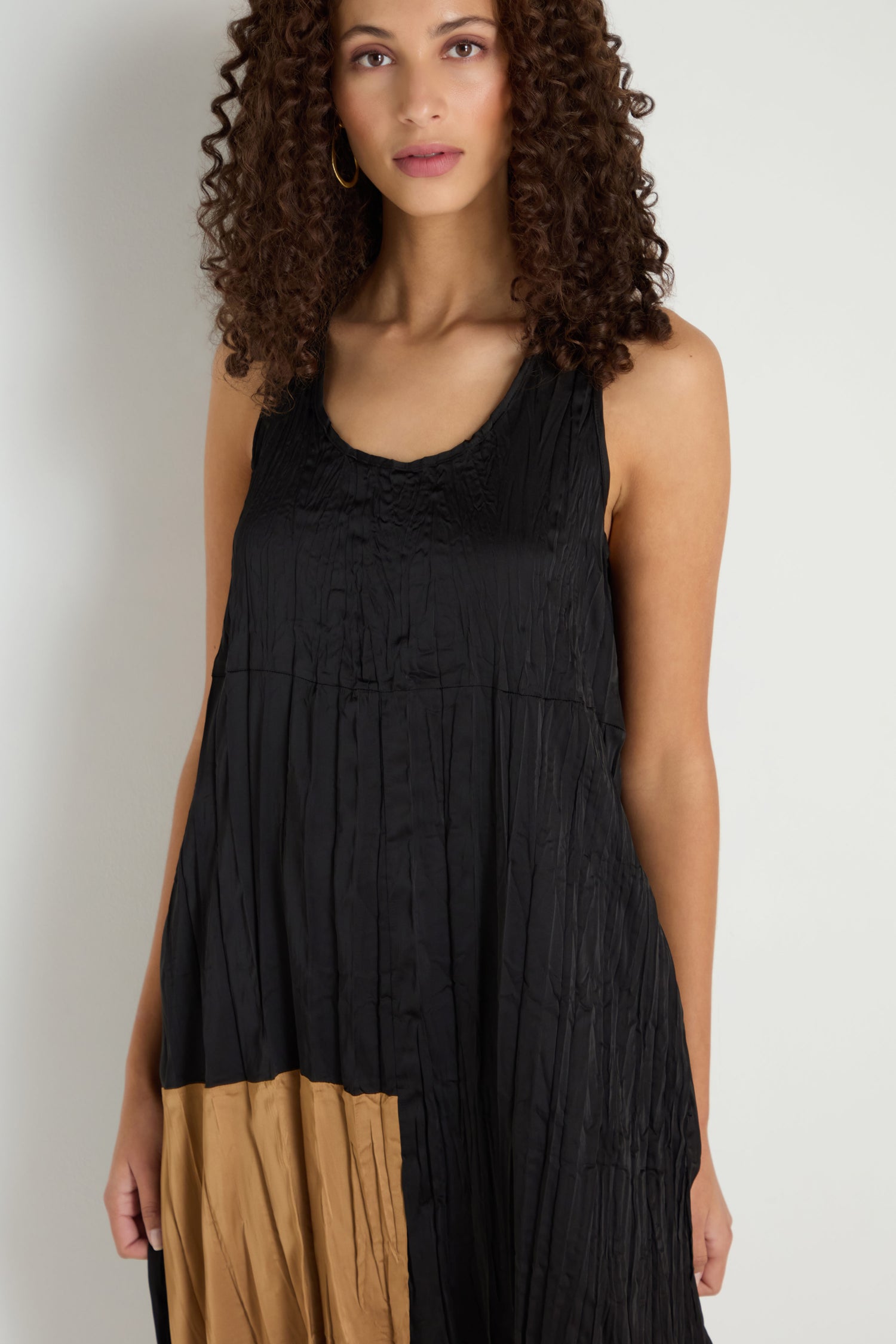 A woman with curly hair models the Colour Block Pleated Dress, a sleeveless black dress featuring a tan block near the hem, set against a plain white background.