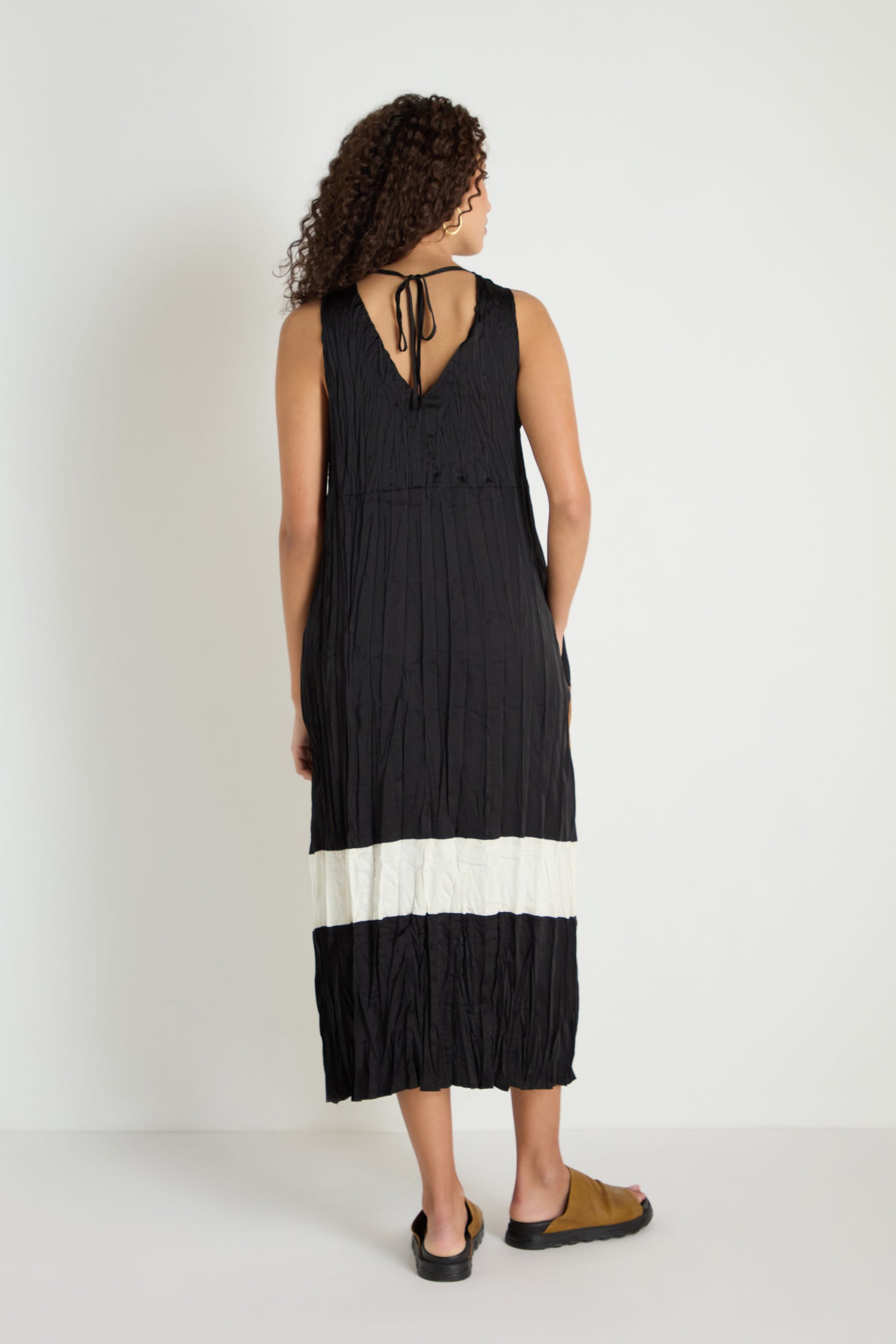 A woman faces away, wearing the Colour Block Pleated Dress—a sleeveless black midi with a white horizontal stripe near the hem—and brown sandals, set against a plain white background.