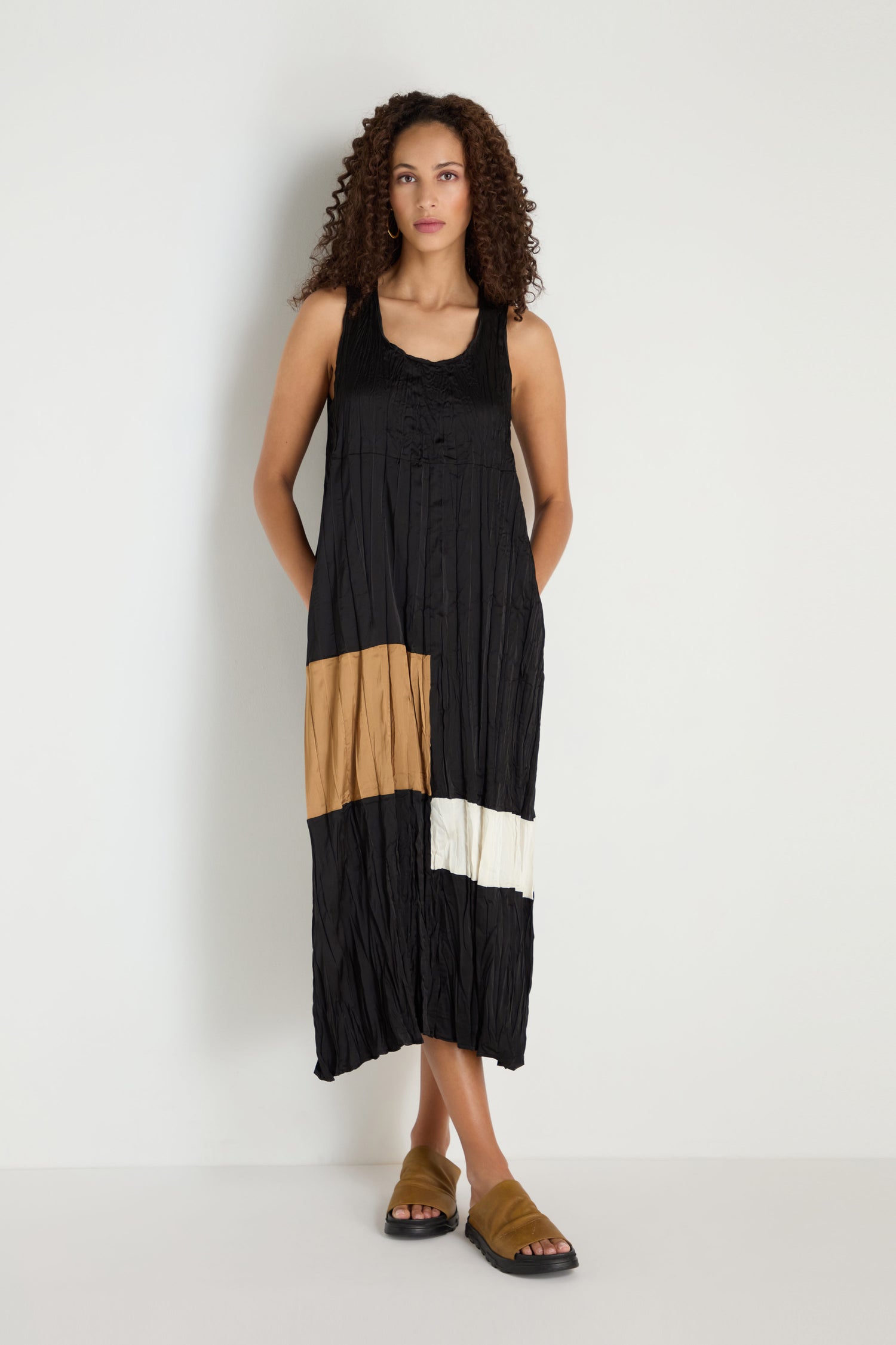 A woman stands against a white wall wearing the Colour Block Pleated Dress, a sleeveless black dress featuring tan and white accents, styled with brown sandals.