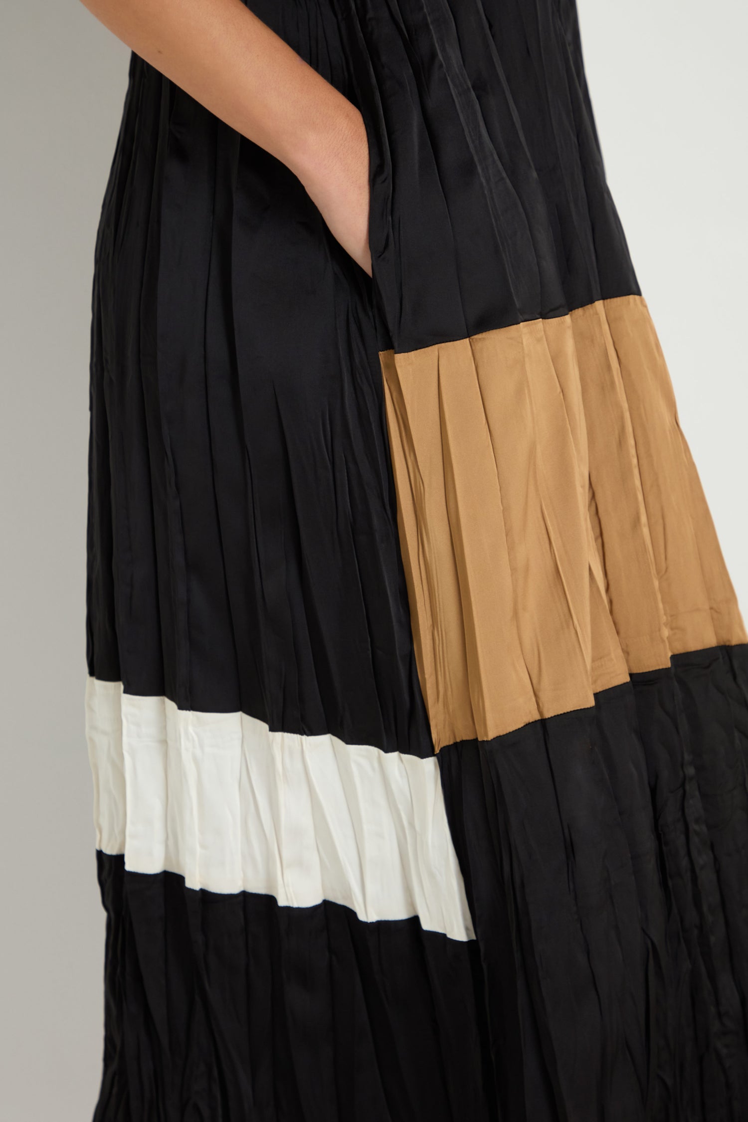 A close-up of the Colour Block Pleated Dress—a black pleated midi featuring beige and white panels—shows a person's hand tucked into the side pocket.