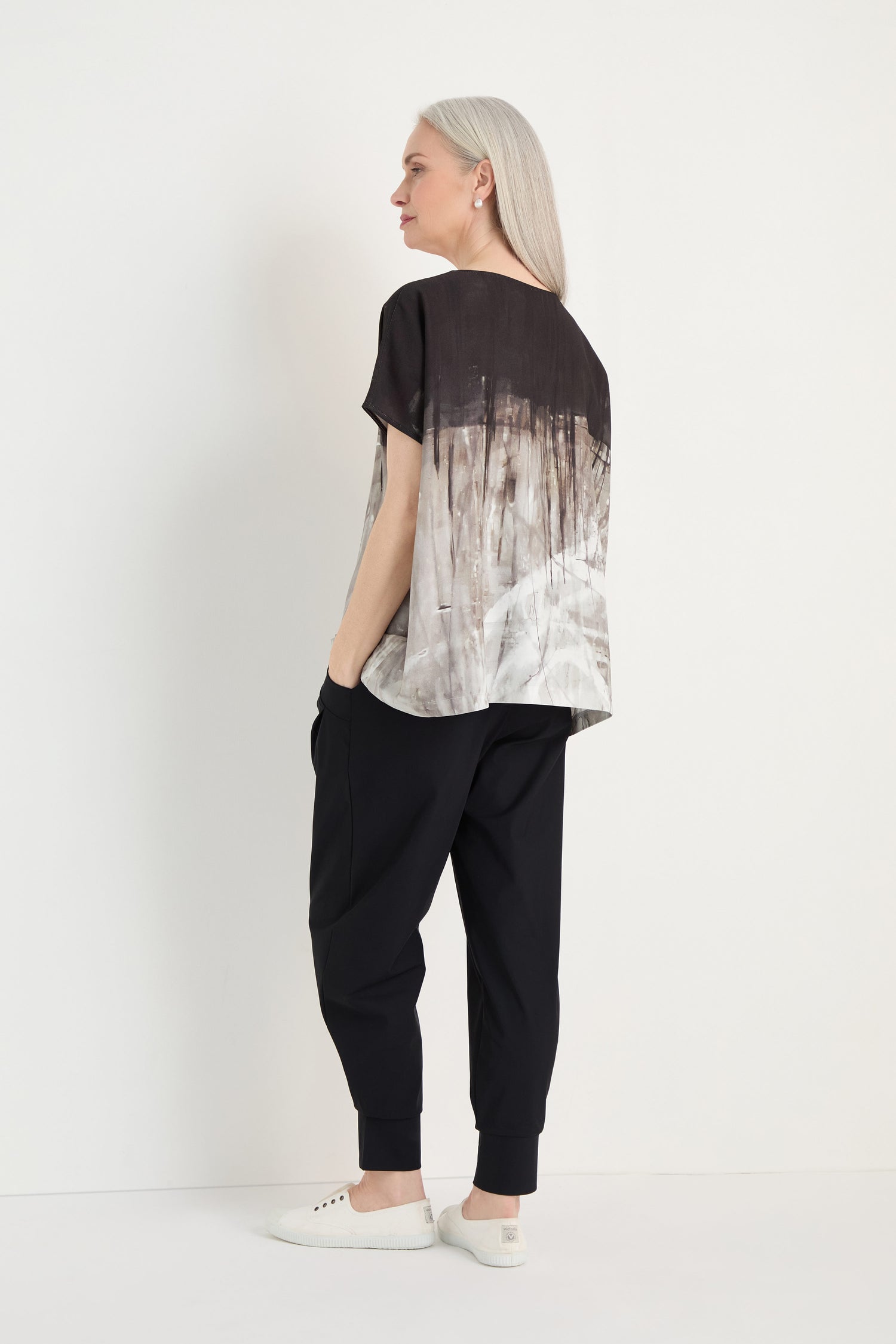 A woman with long gray hair stands sideways against a white background, wearing the Abstract Print Easy Top—a relaxed fit blouse with a monochrome abstract print—paired with black pants and white slip-on shoes.