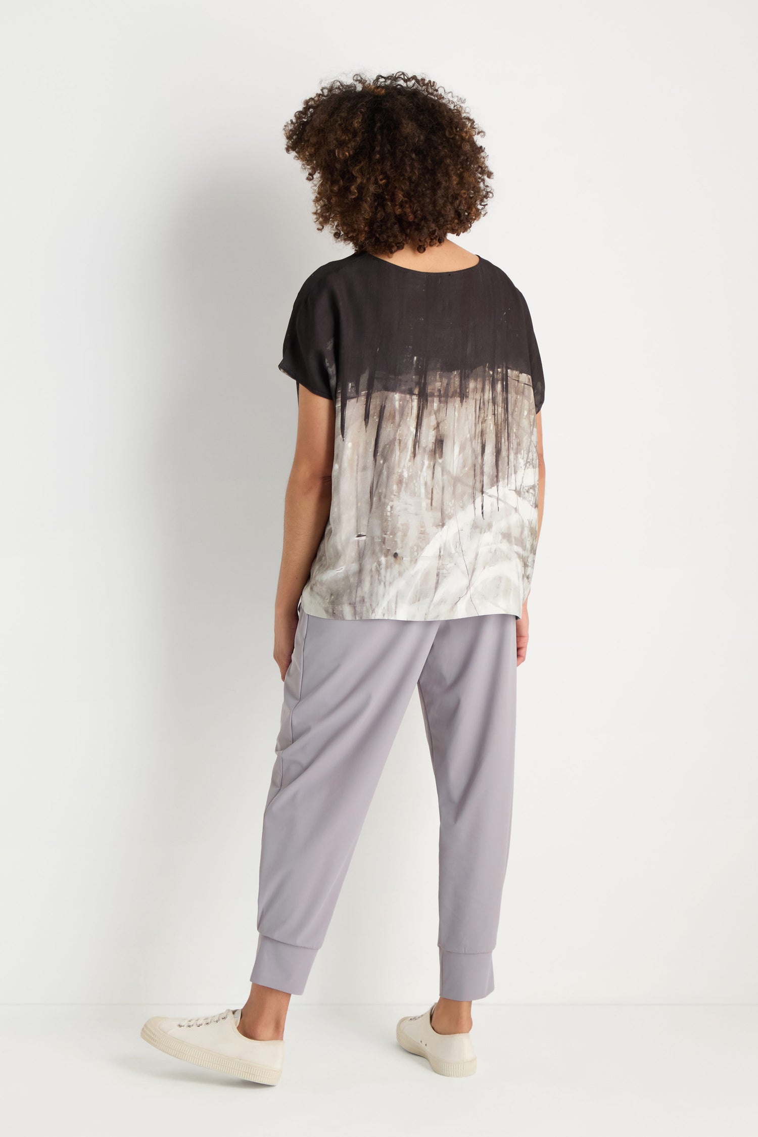 A woman with long gray hair stands sideways against a white background, wearing the Abstract Print Easy Top—a relaxed fit blouse with a monochrome abstract print—paired with black pants and white slip-on shoes.