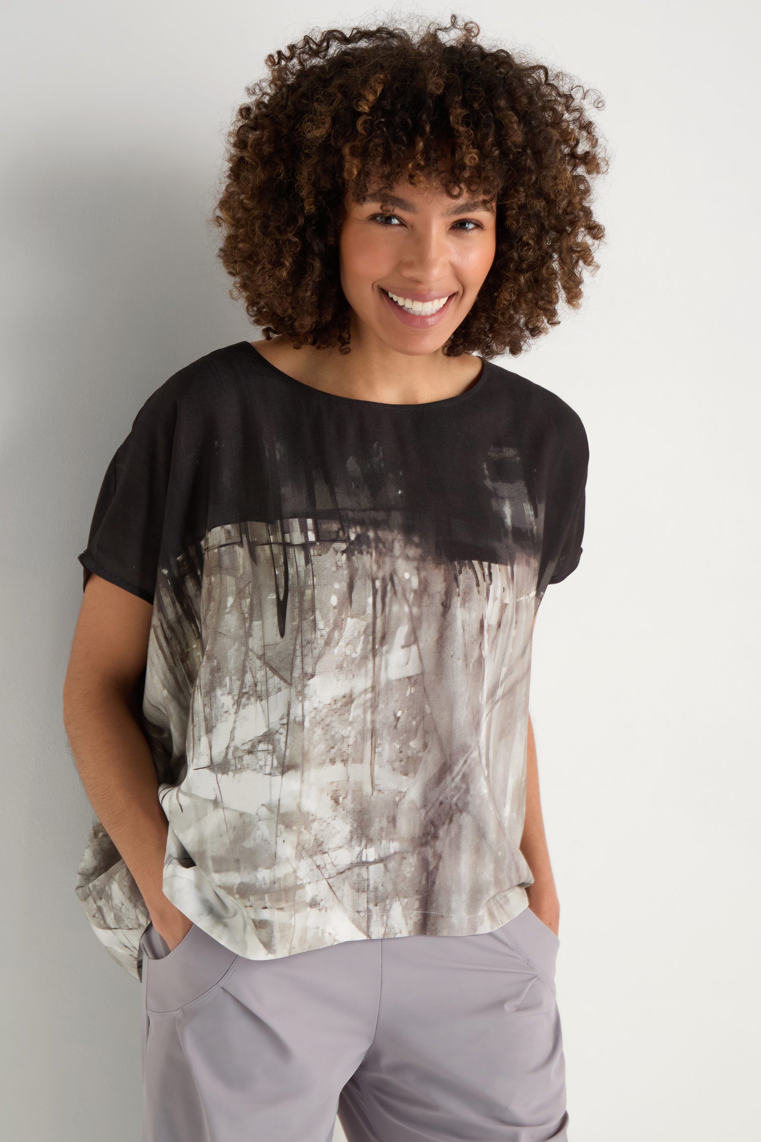 A woman with long gray hair wears the Abstract Print Easy Top in black and gray, paired with black pants, standing with her hands in her pockets against a plain background.