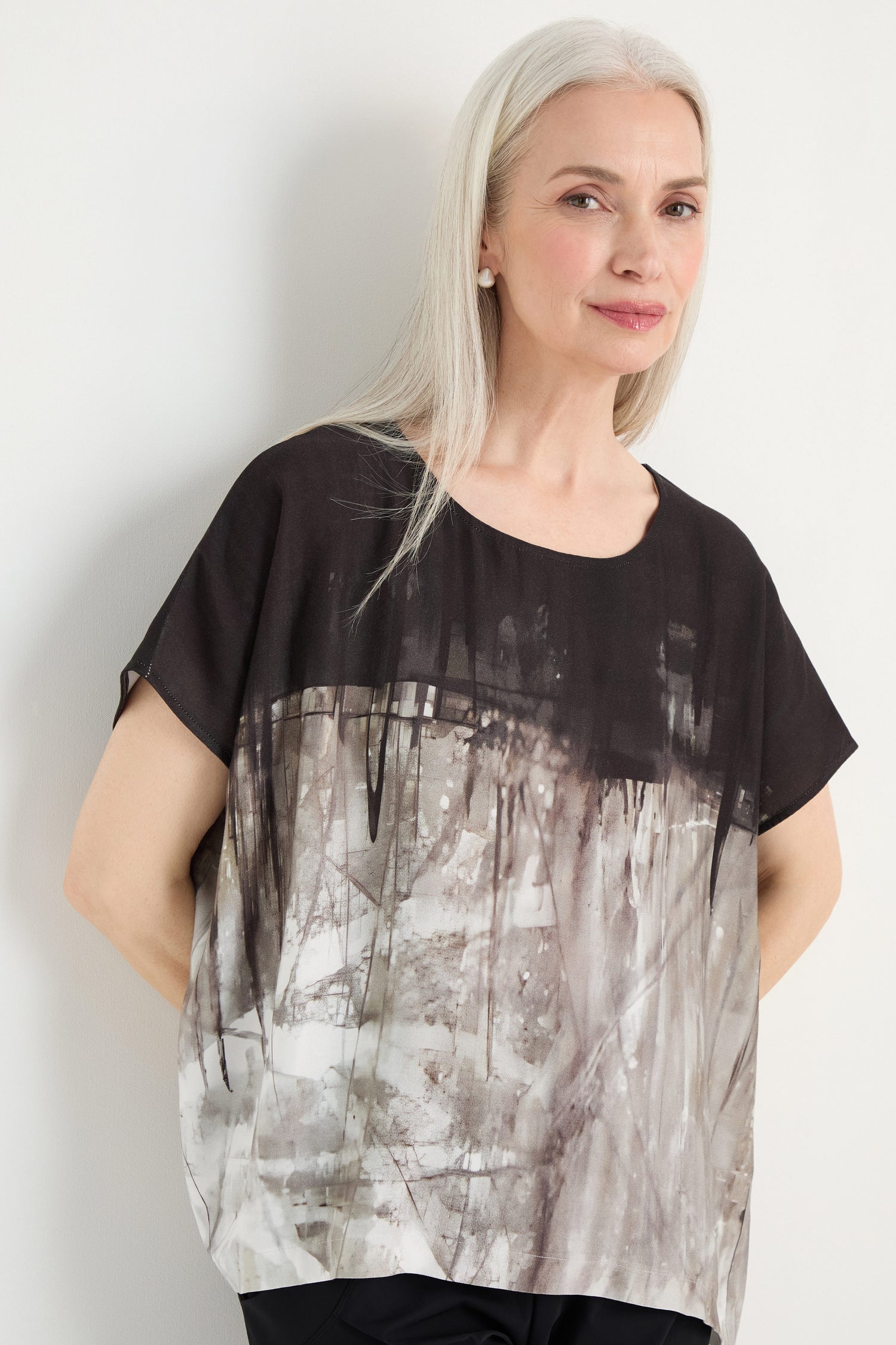 A woman with long gray hair stands against a light background wearing the Abstract Print Easy Top, a relaxed-fit blouse featuring an abstract black and gray pattern.