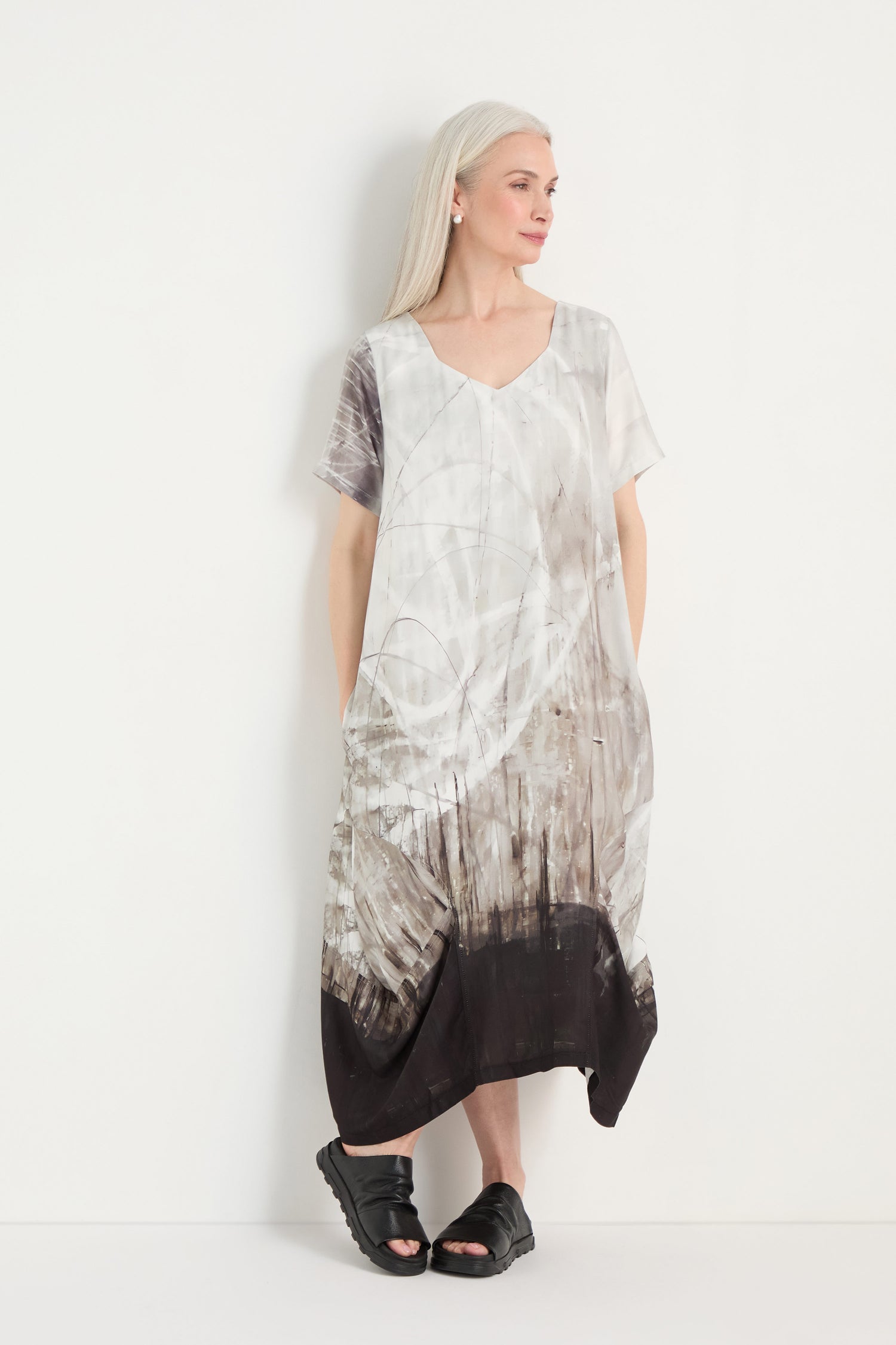 A woman with long white hair stands against a white background, wearing the Abstract Print Bubble Dress with an abstract gray and black pattern and black sandals.