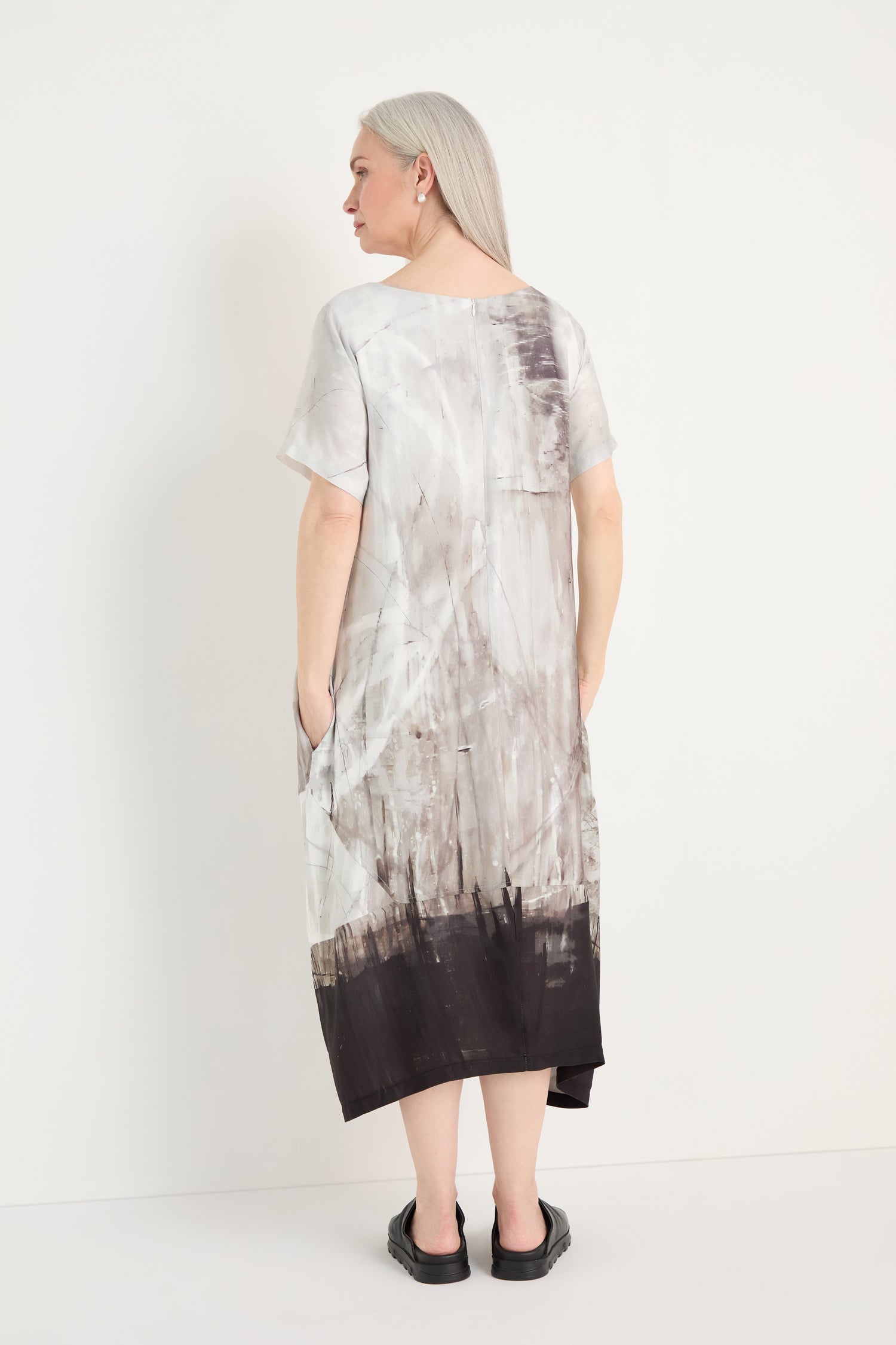 A person with long gray hair stands facing away against a plain white background, wearing a relaxed fit, short-sleeve Abstract Print Bubble Dress and black slip-on shoes.