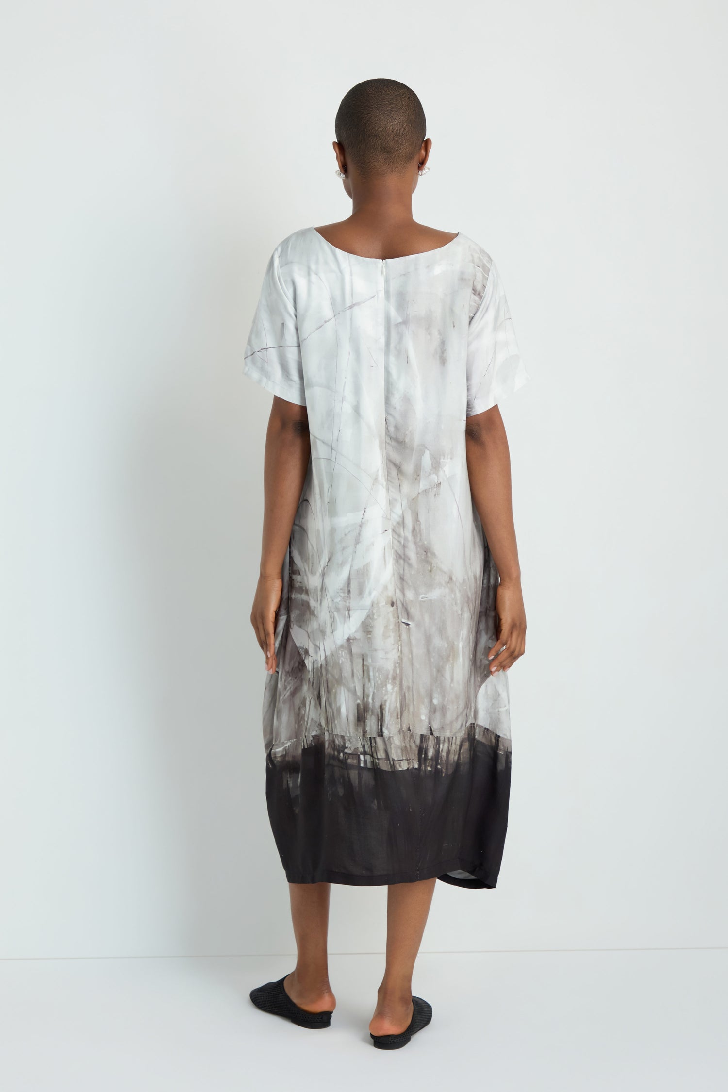 A person with long gray hair stands facing away against a plain white background, wearing a relaxed fit, short-sleeve Abstract Print Bubble Dress and black slip-on shoes.