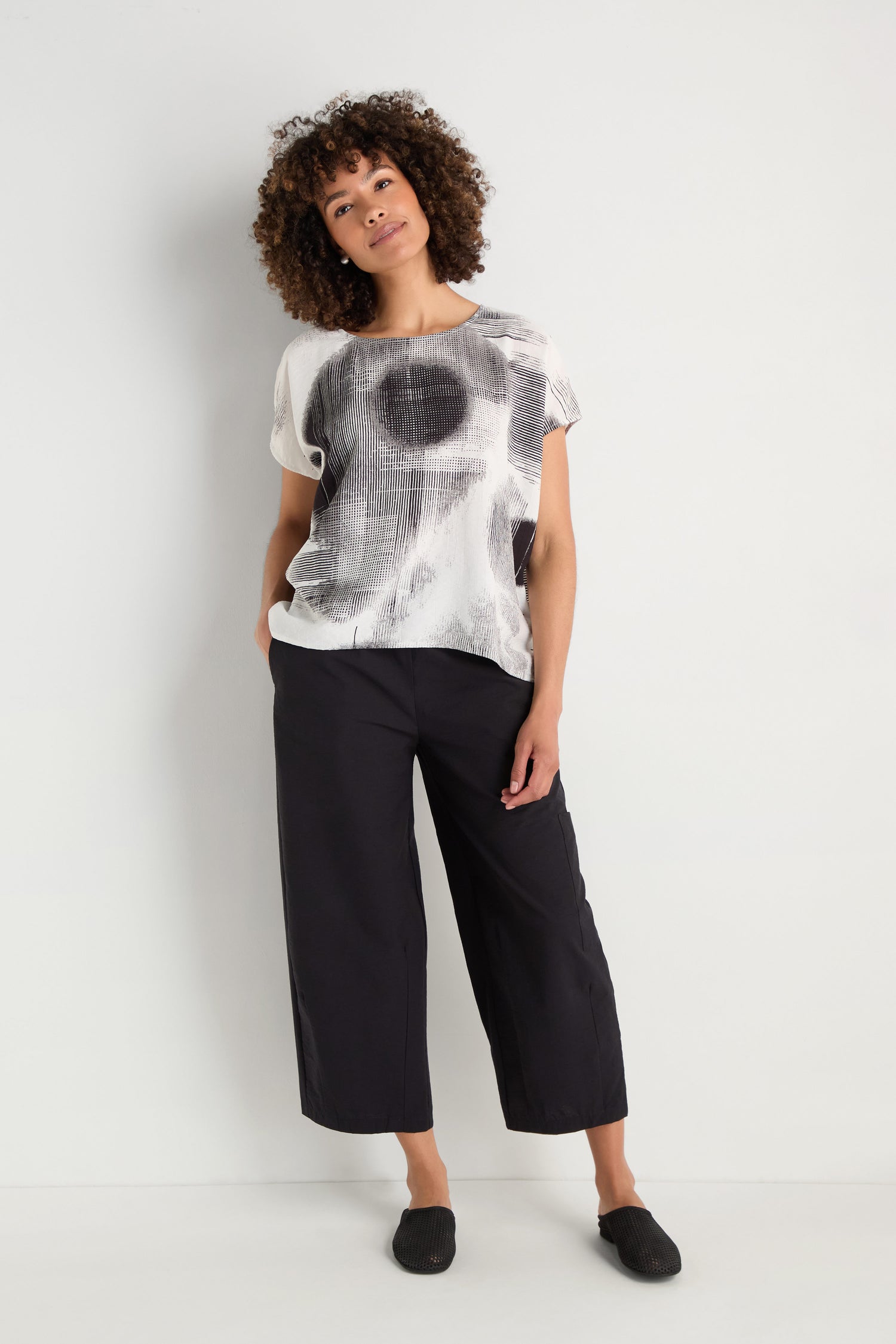 A woman stands against a white wall wearing a Shaded Print Top with a relaxed silhouette, light grey tapered pants, and white sneakers.