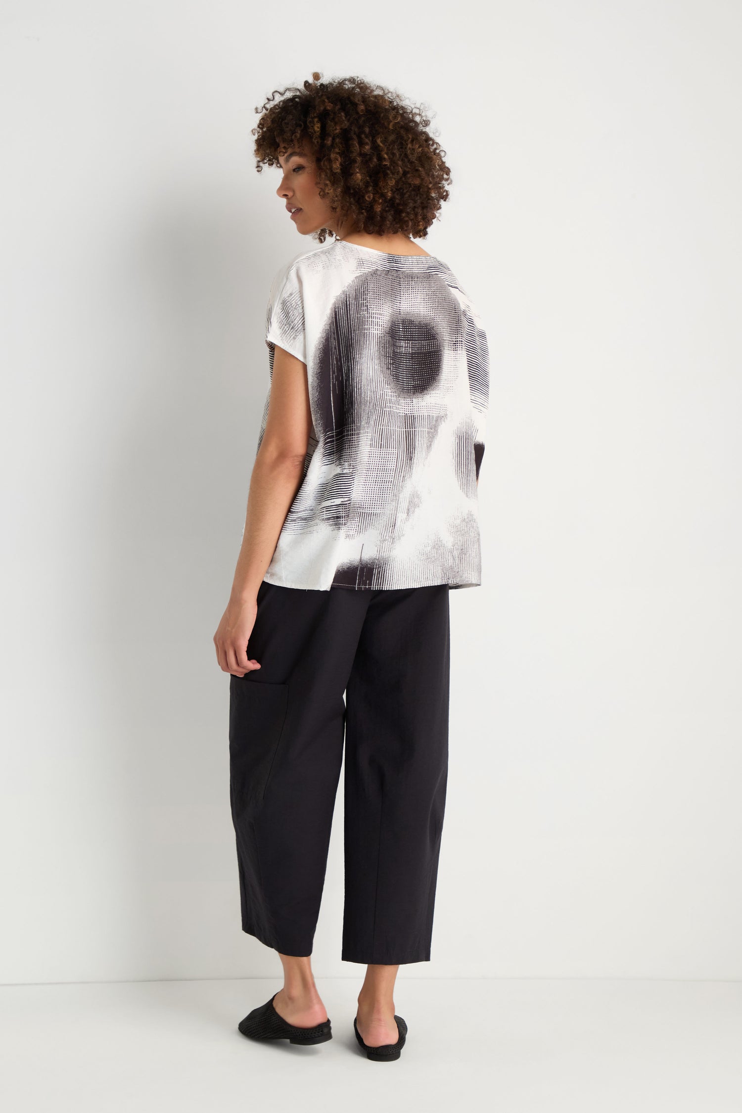 A person with long gray hair faces a white wall, wearing the Shaded Print Top with a relaxed silhouette, gray pants, and white shoes.