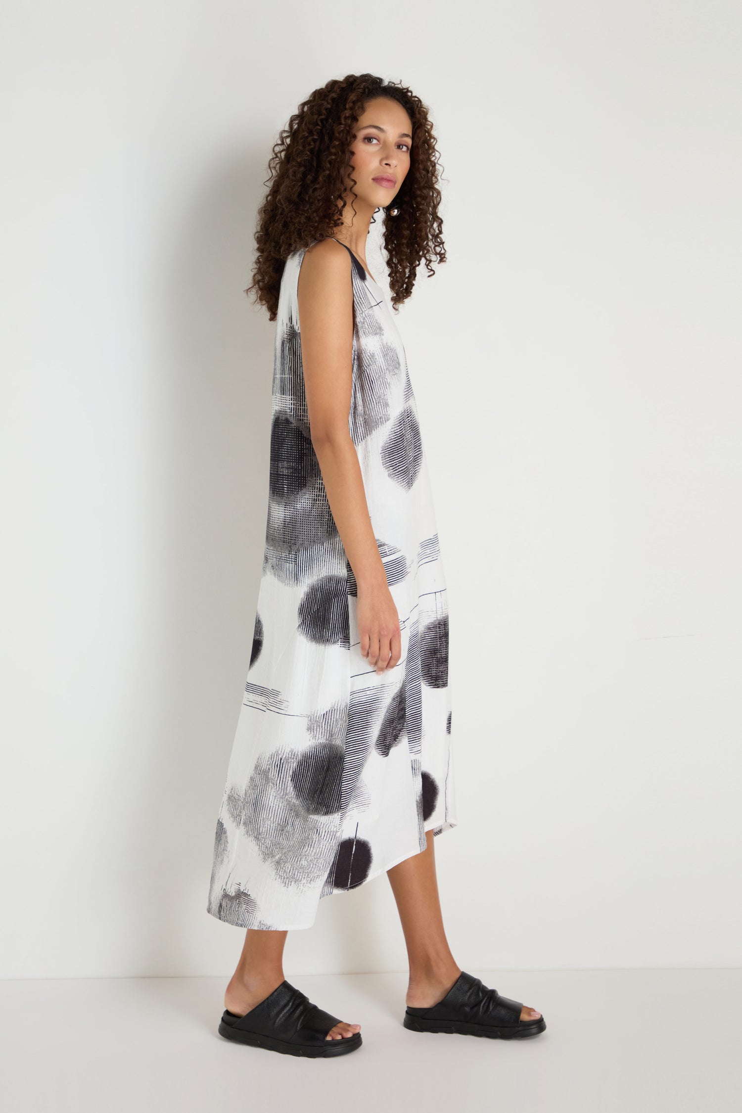 A woman with curly hair wears the Shaded Print Flared Dress—sleeveless, loose-fitting, monochrome with black abstract patterns—paired with black sandals, standing against a plain white background.