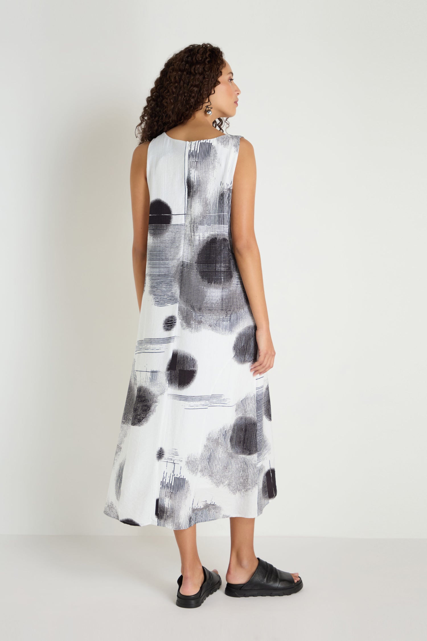 A woman faces away, wearing the Shaded Print Flared Dress with black abstract spots and patterns, plus black sandals, set against a plain light background.