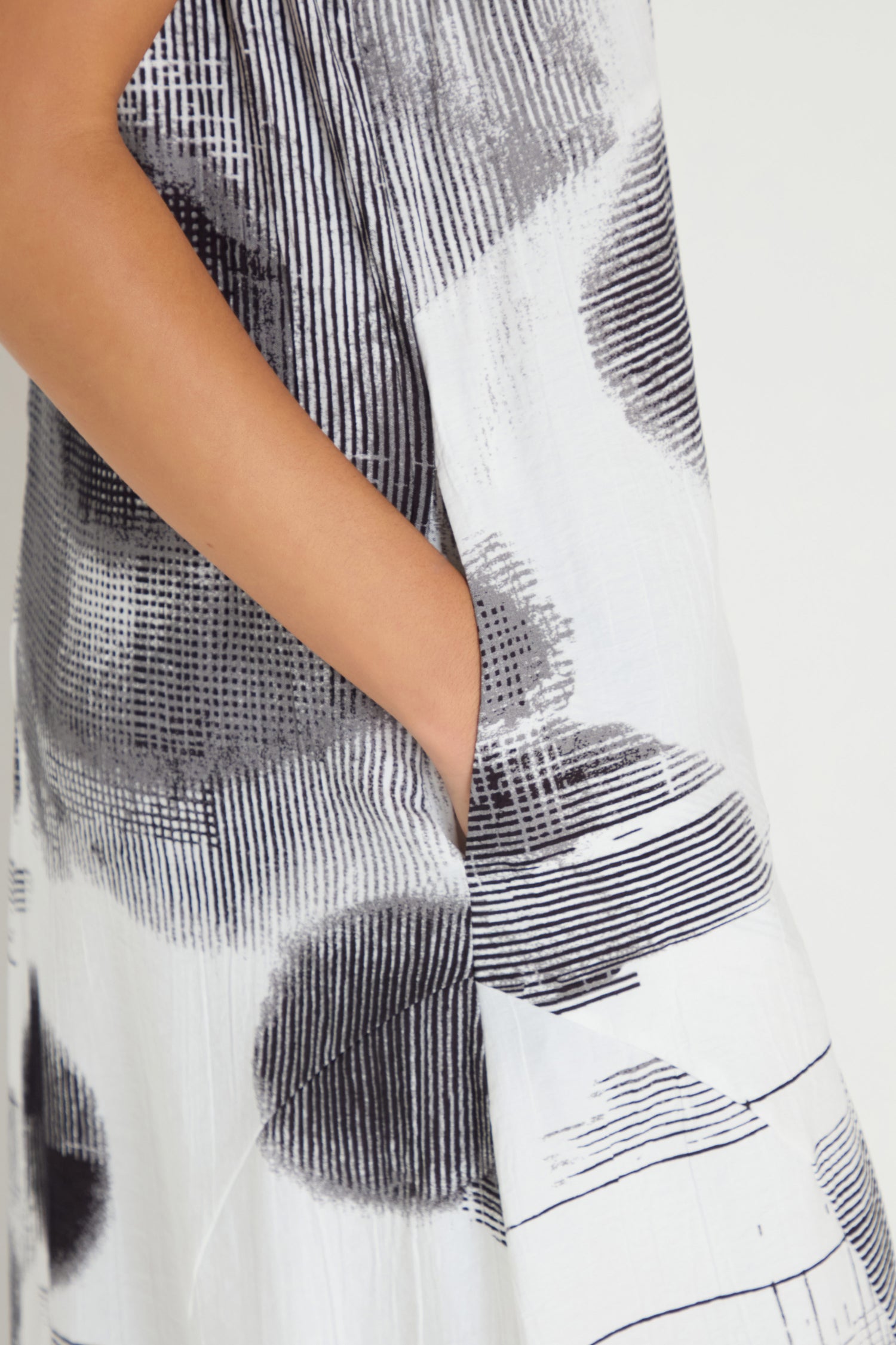 A person wears the Shaded Print Flared Dress, a sleeveless midi outfit featuring abstract black patterns and side pockets, with one hand in a pocket.