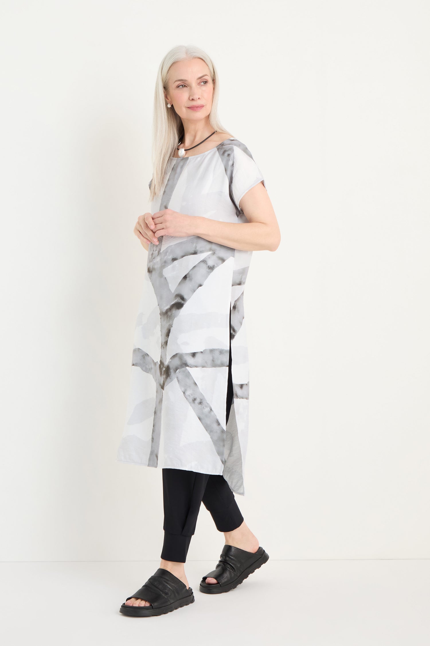 A woman with long white hair models the Brushstroke Print Long Tunic, featuring short sleeves and a knee-length hem, styled with black leggings and sandals against a plain white background.
