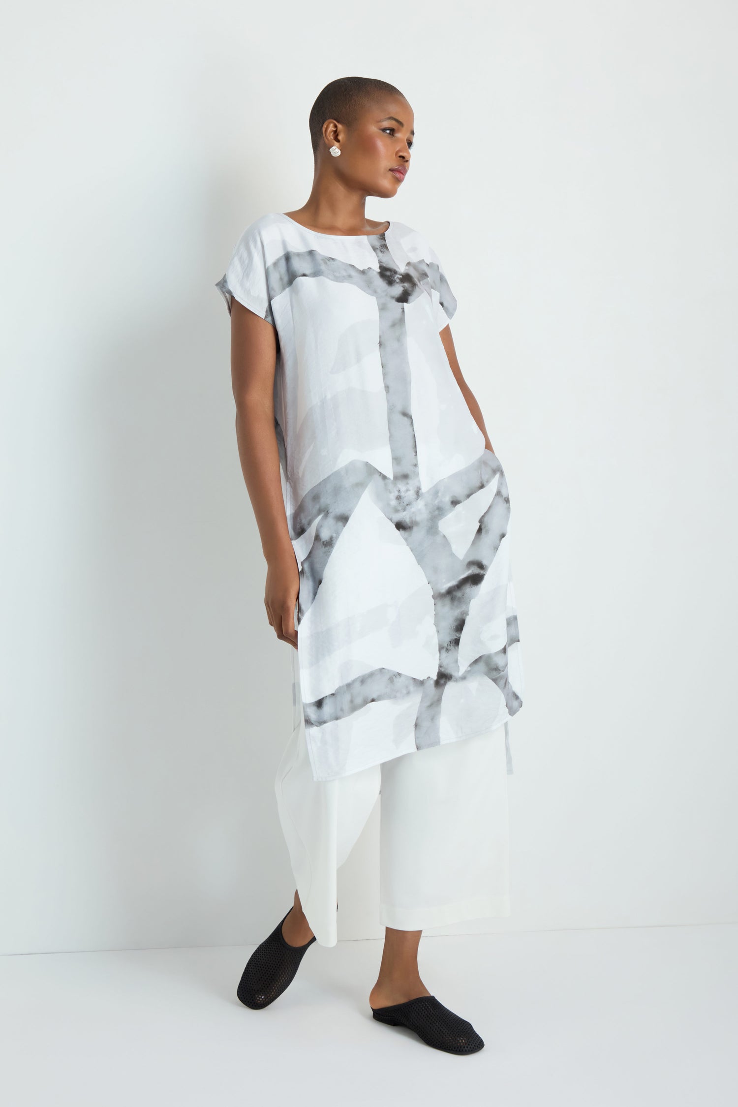 A woman with long white hair models the Brushstroke Print Long Tunic, featuring short sleeves and a knee-length hem, styled with black leggings and sandals against a plain white background.