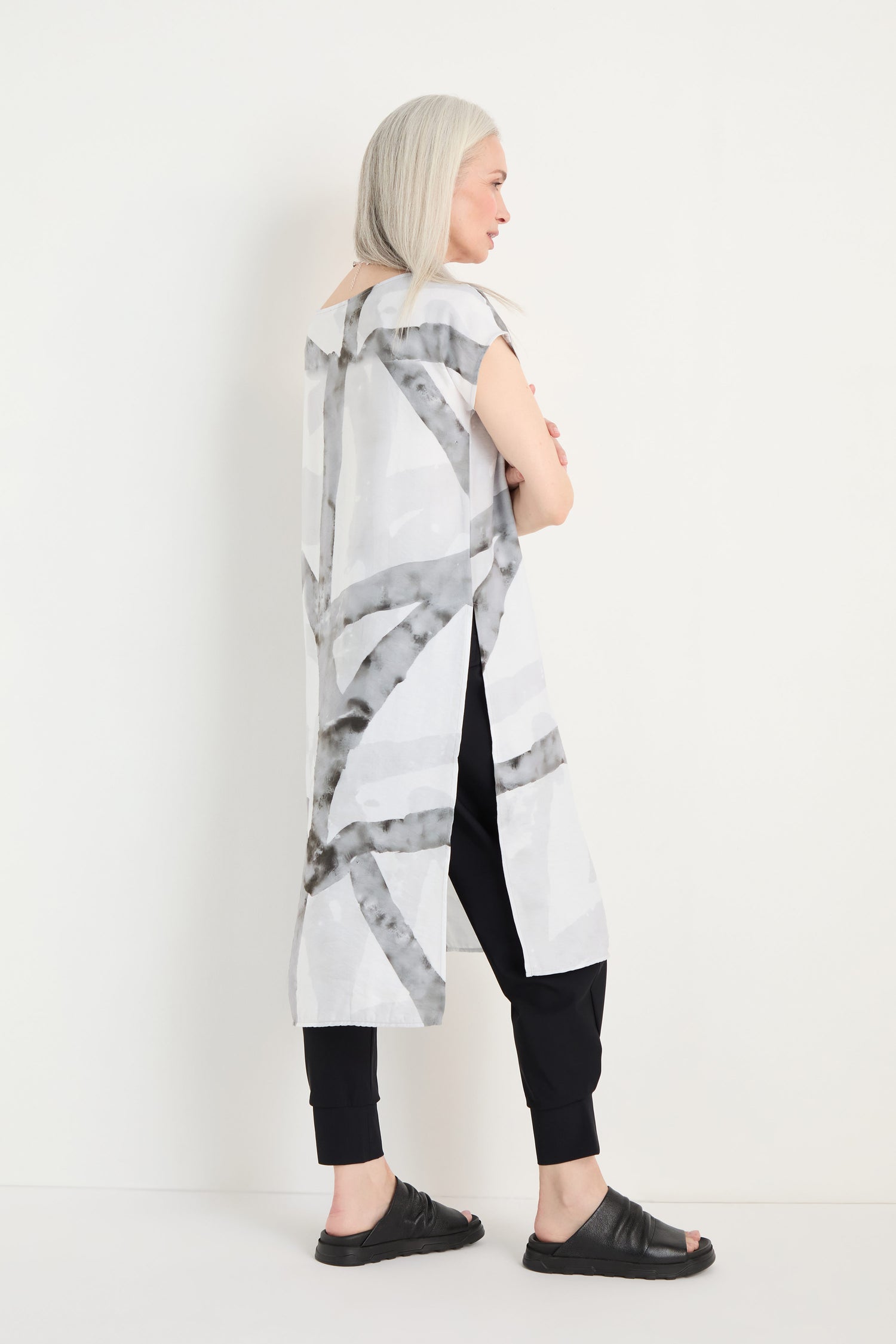 A woman with long gray hair wears the Brushstroke Print Long Tunic over black pants and sandals, standing sideways against a plain white background.