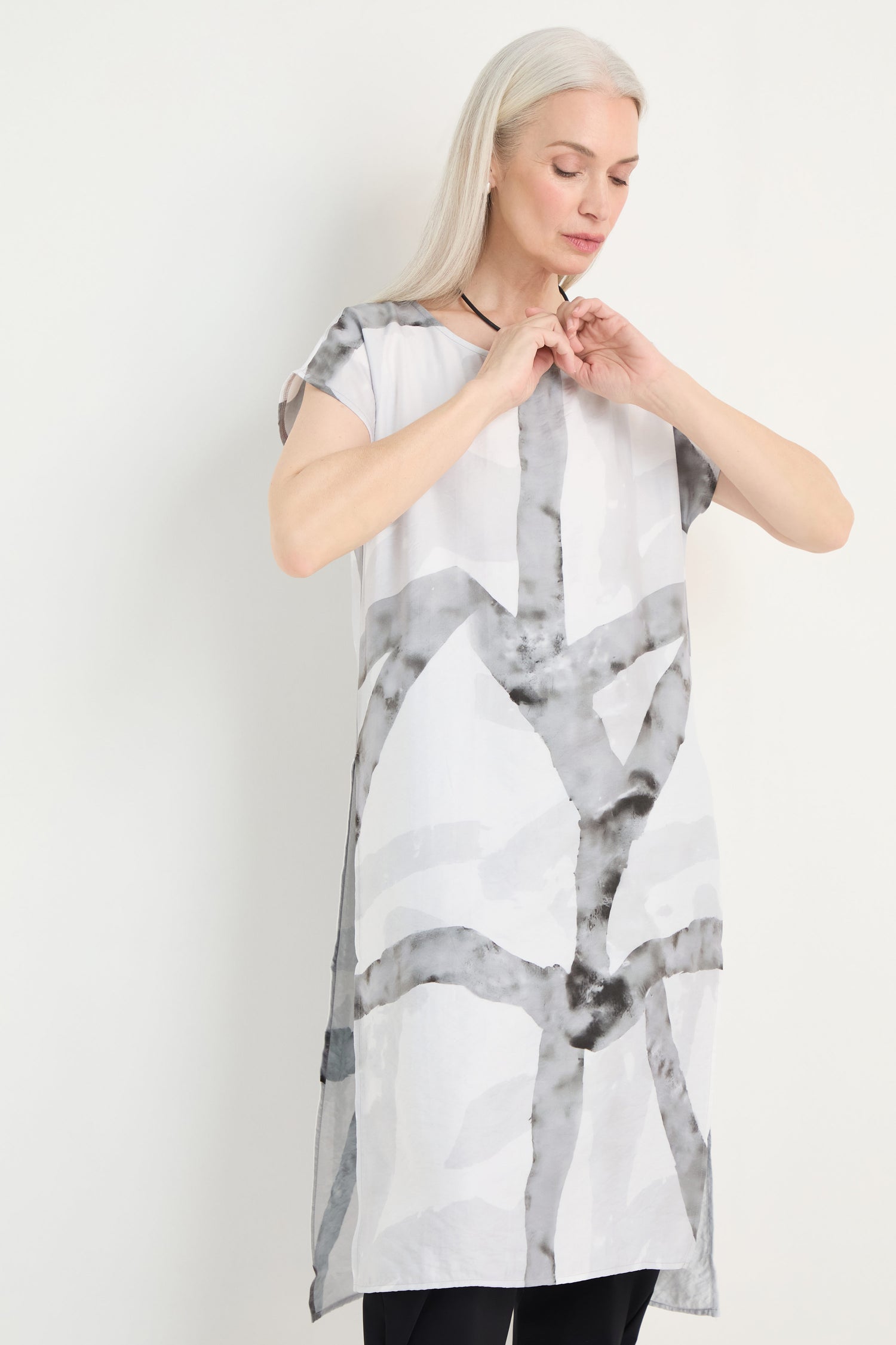 A woman with long gray hair wears the Brushstroke Print Long Tunic, featuring a loose fit and abstract gray patterns. She stands against a plain background, looking down as she adjusts her necklace.