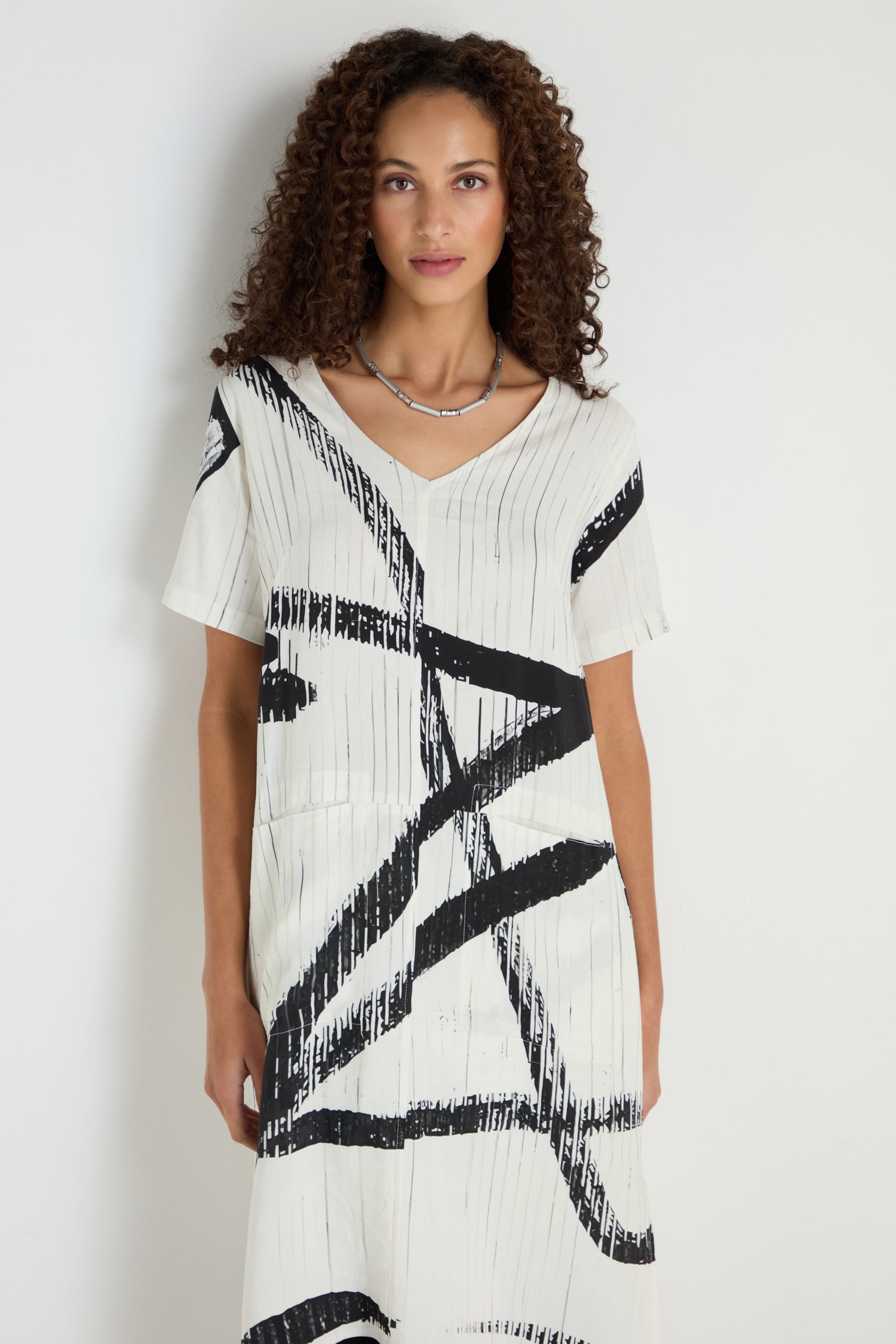 Woman with curly hair models the Soft Stripe Midi Dress—a relaxed midi silhouette in white viscose-linen blend with bold black stripes—set against a plain white background.
