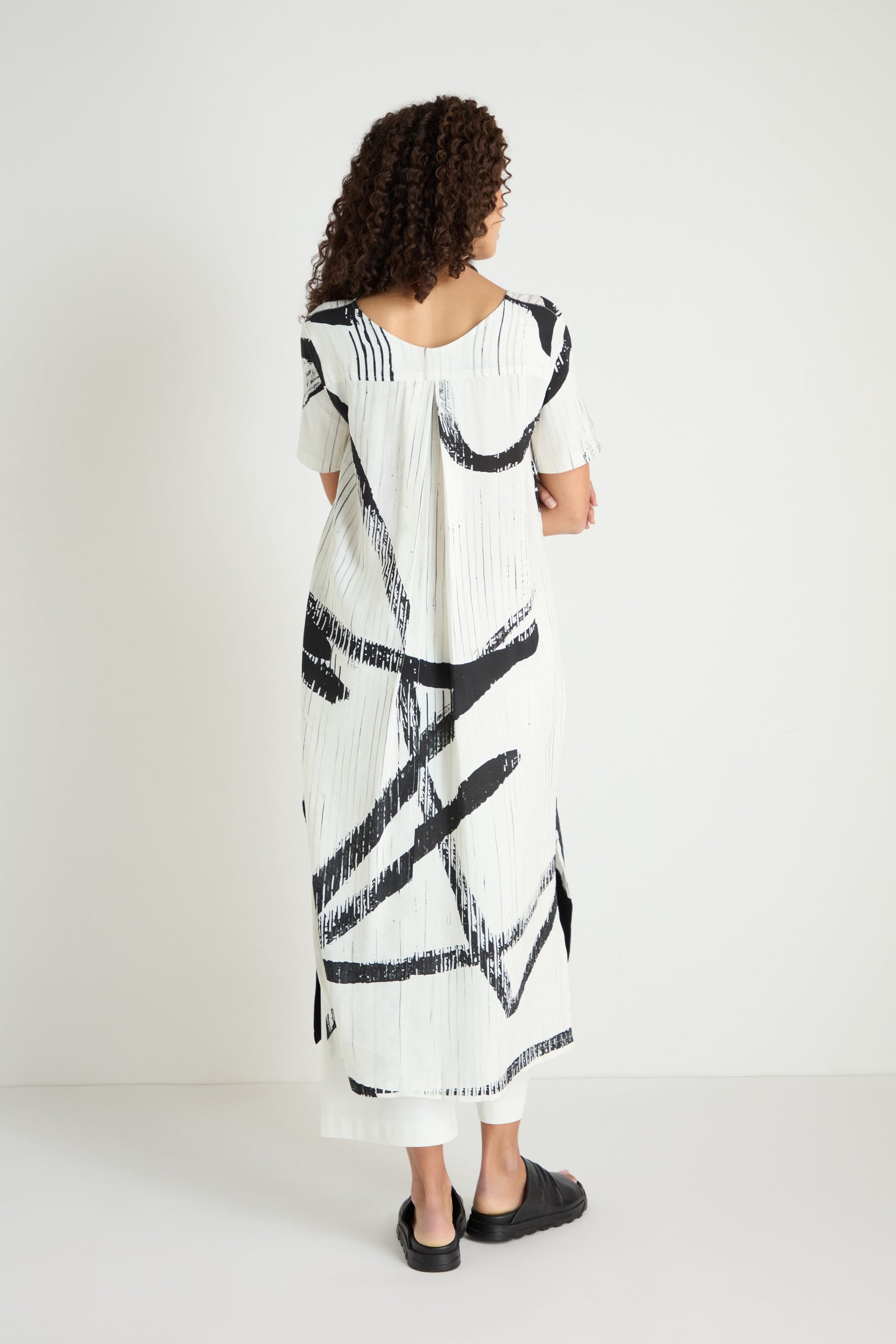 A person with curly hair, facing away, wears the Soft Stripe Midi Dress—a relaxed viscose linen blend with bold black abstract patterns—and black sandals, set against a plain white background.