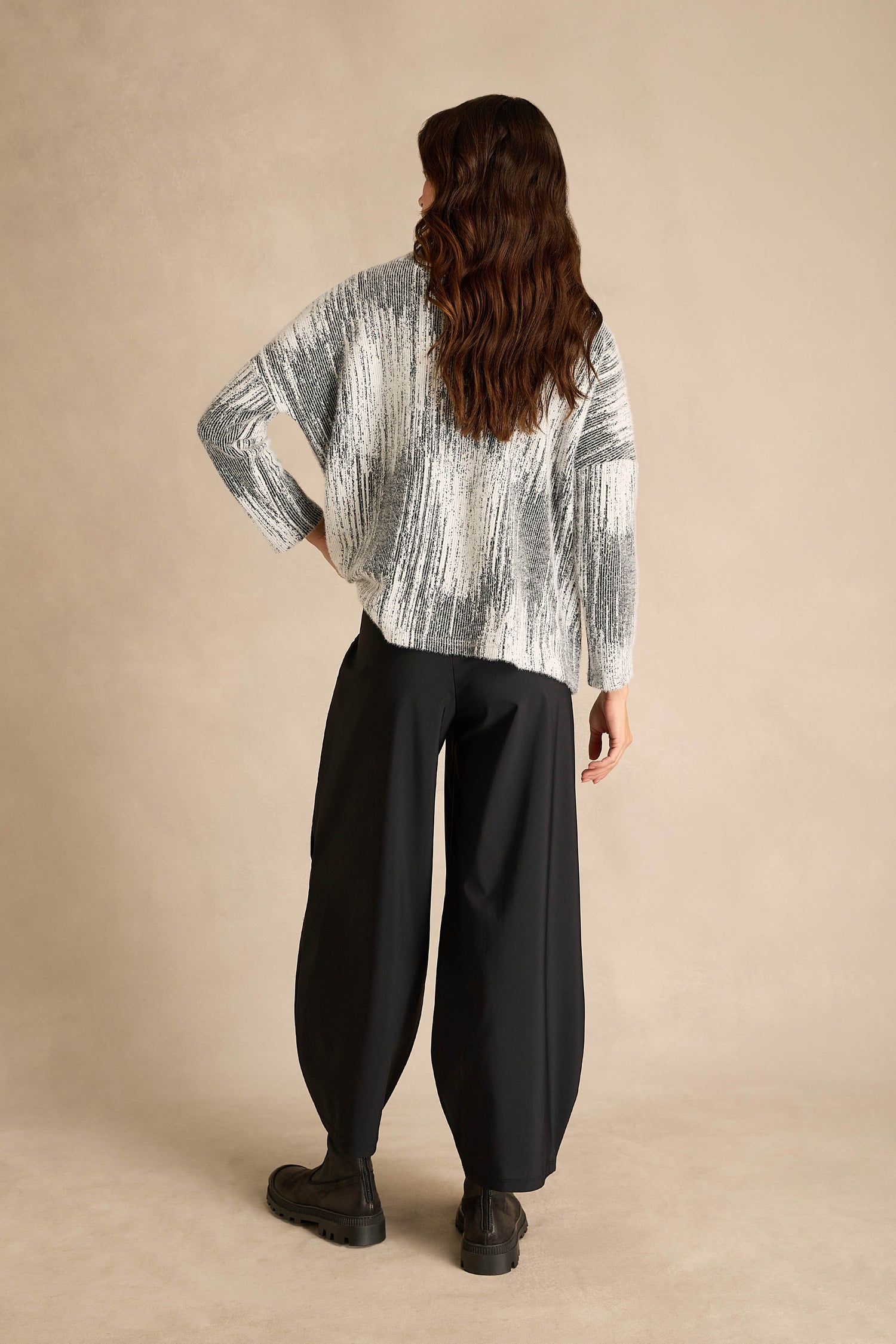 A person stands with their back to the camera, wearing a black and white patterned sweater, Seamed Bubble Trousers in a wide-leg stretch fabric, and black boots, set against a plain beige background.