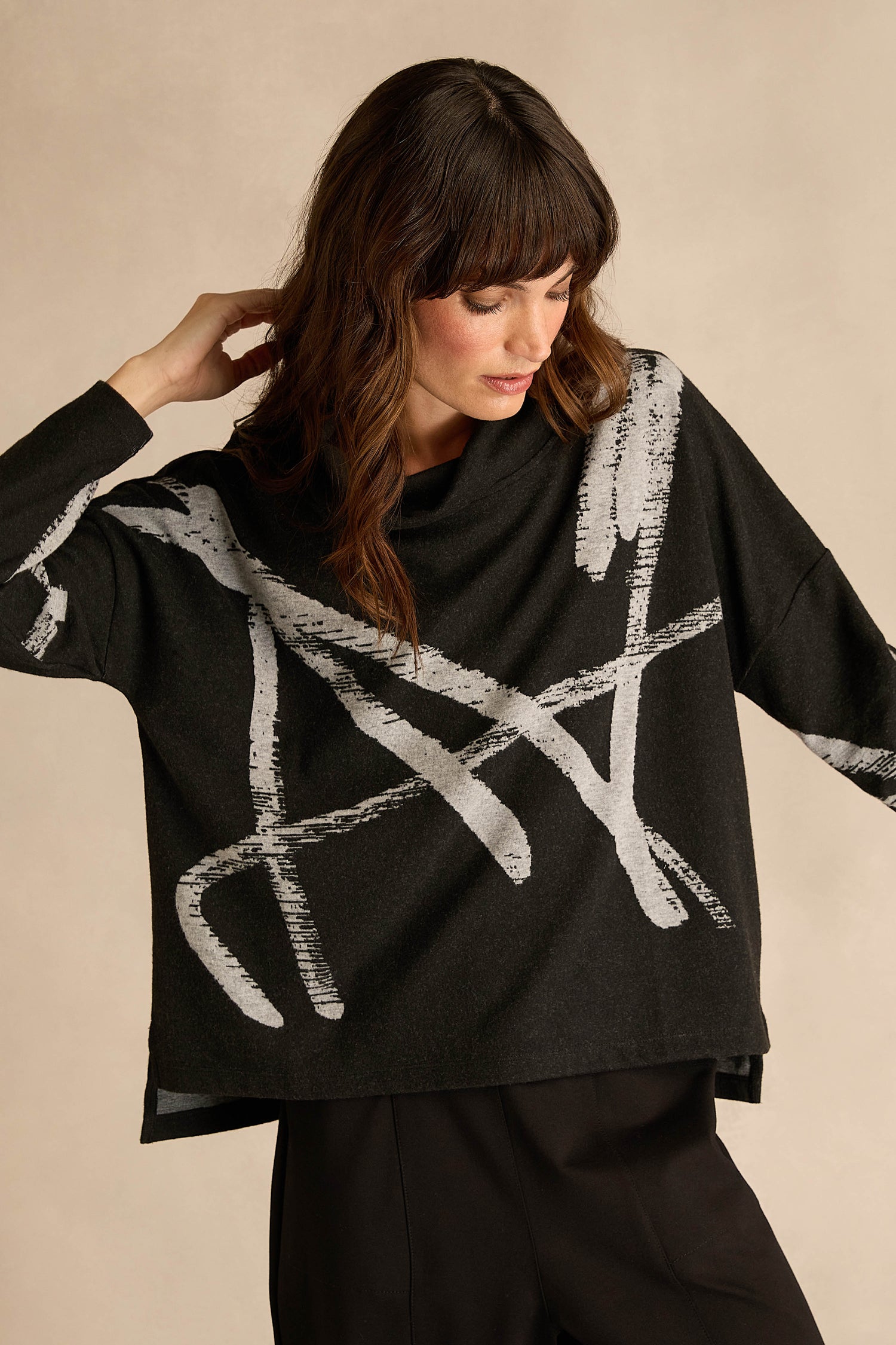 A woman wearing the Paint Stripes Long Sleeve Top—a loose black jersey featuring bold white abstract brushstroke patterns—stands against a plain beige background.