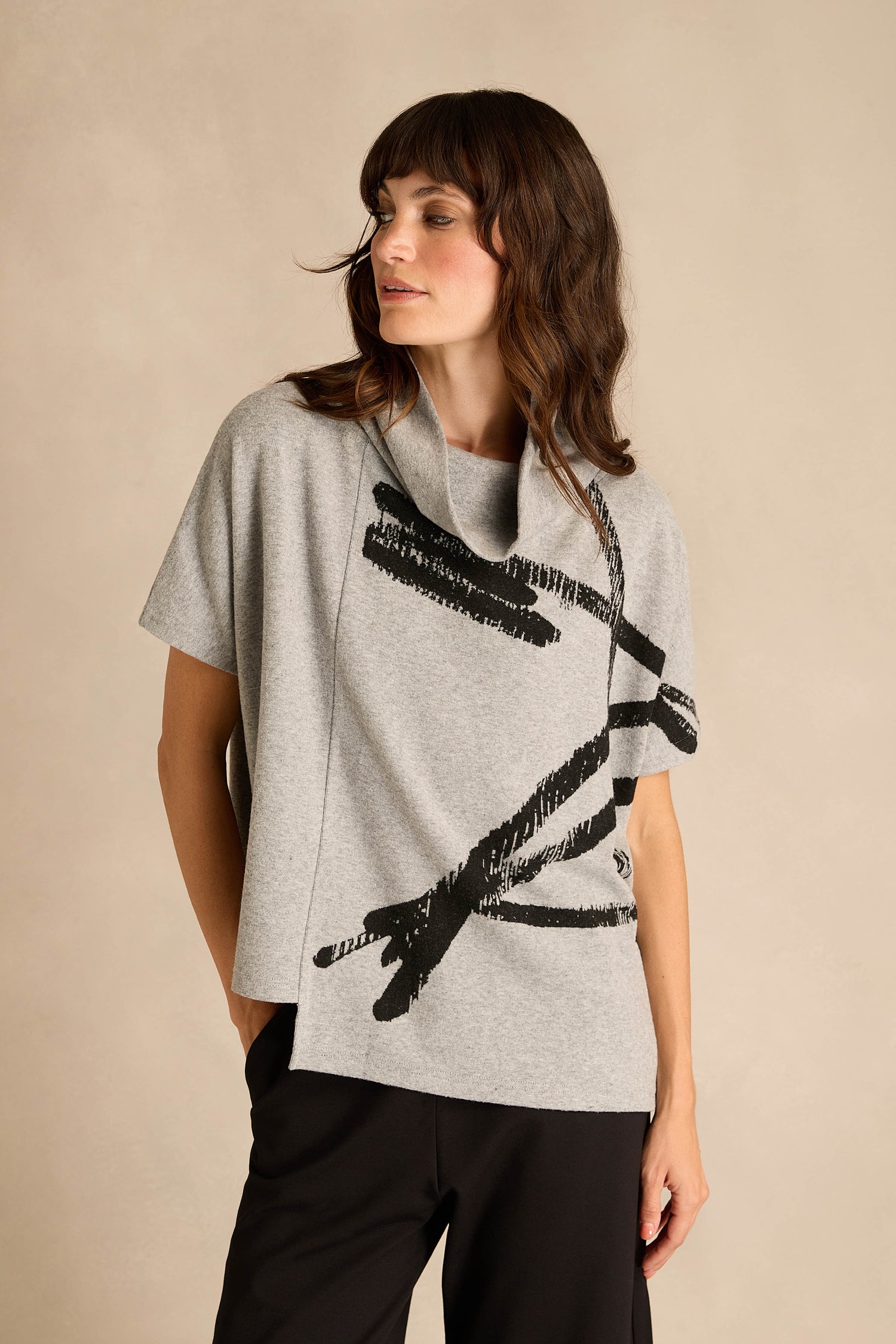 Woman wearing the Paint Stripes Draped Neck Top, a short-sleeved gray top with a black abstract brushstroke design, paired with black pants and standing against a plain beige background.