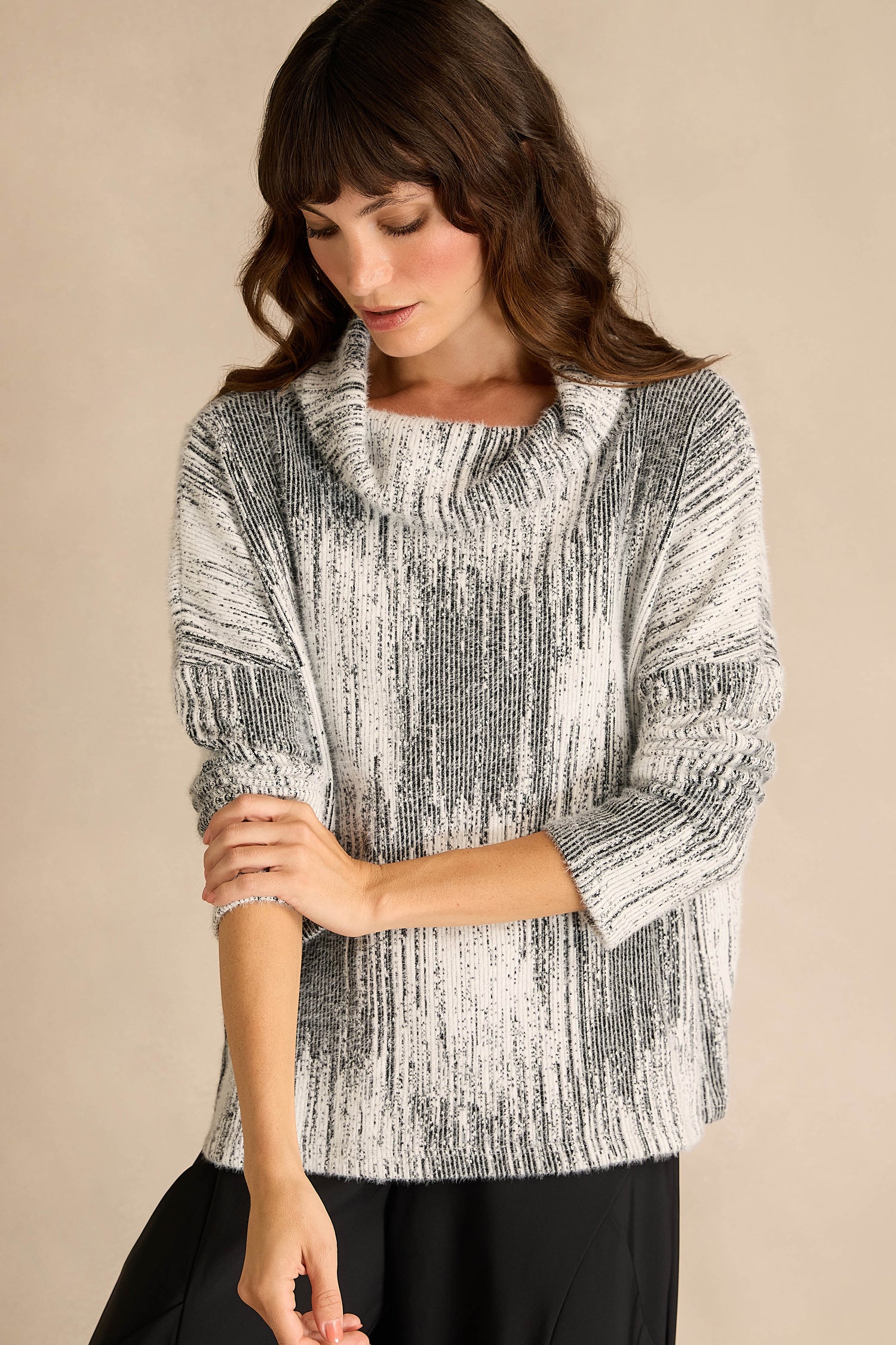 A woman wearing the Greyscale Textured Jersey Top—a loose, black-and-white jersey with a subtle textured pattern, long sleeves, and a draped neck—stands against a plain beige background, looking down and touching her arm.